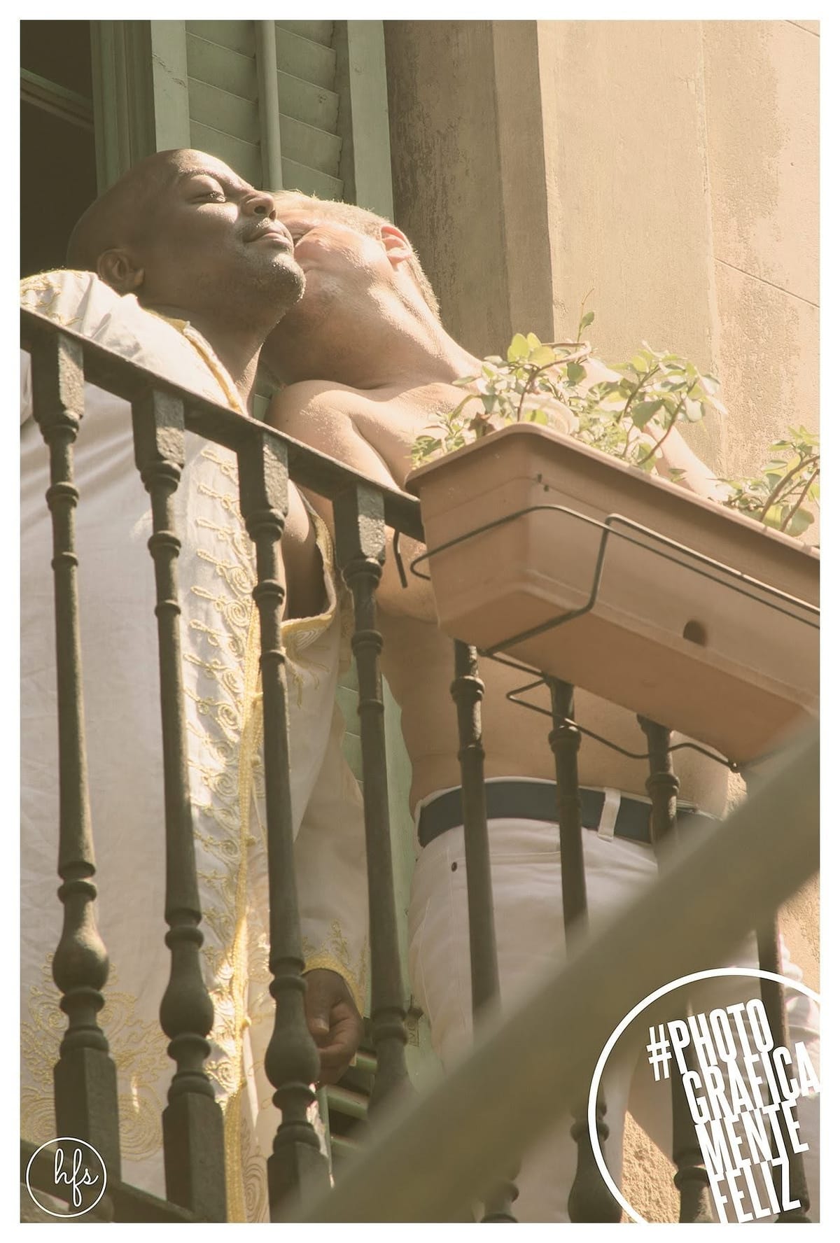 Vista en contrapicado de un edificio de balcones de hierro forjado; en uno de ellos, un joven sin remera se asoma sonriendo y saluda con el brazo extendido, junto a una maceta.