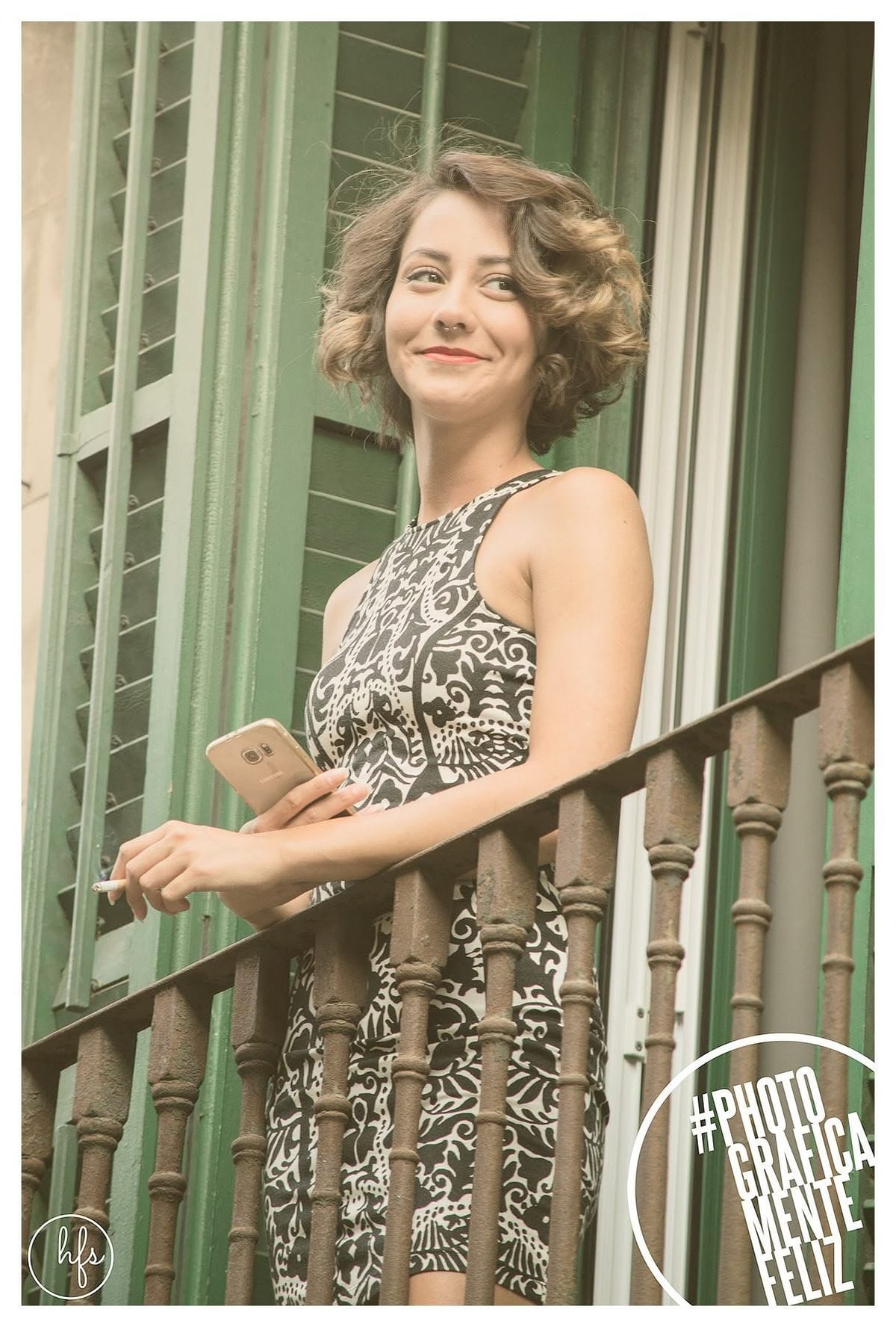 Mujer de perfil sonriente, con pelo rizado y corto, apoyada en la baranda de un balcón con persianas verdes, sosteniendo un celular en una mano y un cigarrillo en la otra.