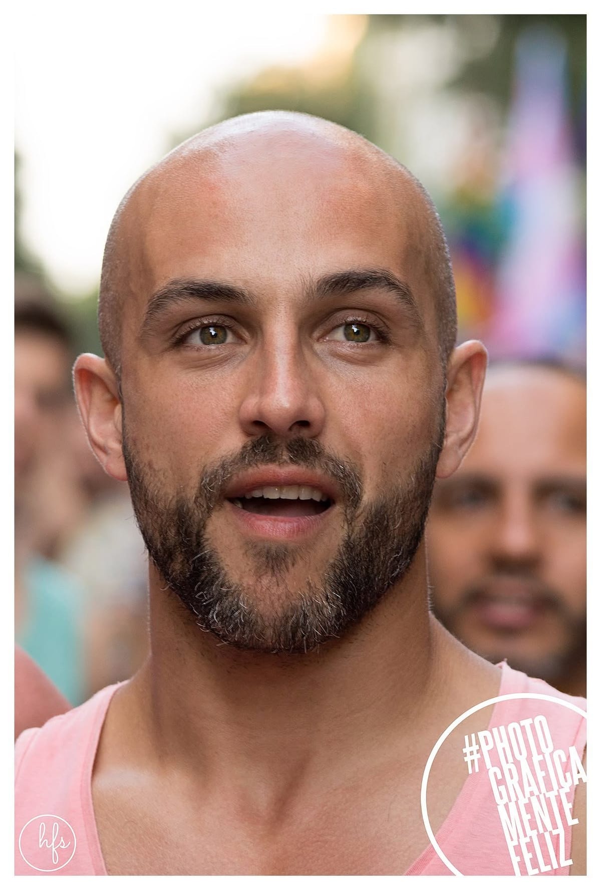 Retrato frontal de un hombre calvo con musculosa rosa, mirada intensa y boca entreabierta, entre otras personas desenfocadas en un evento callejero.