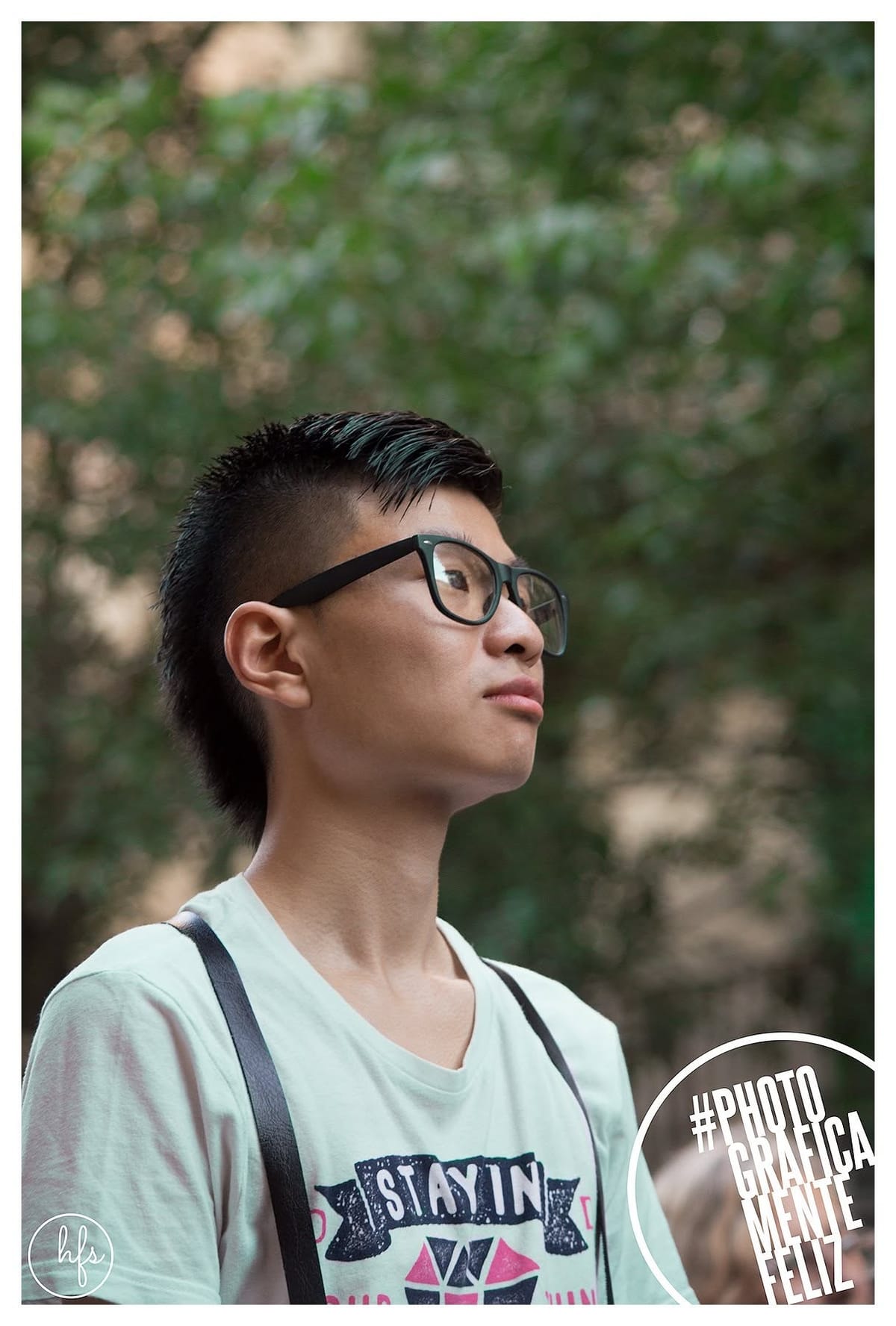 Retrato a color de un joven asiático con anteojos de marco oscuro y corte undercut, mirando hacia arriba a la derecha; viste una remera clara con estampado "STAYIN'" y tirantes de mochila…