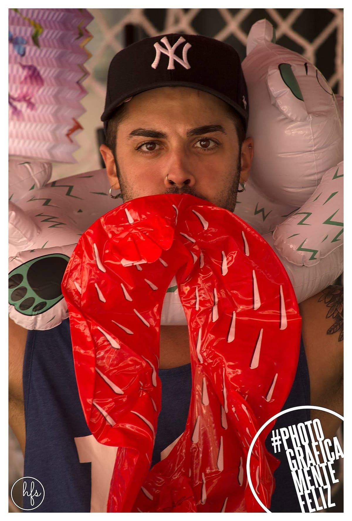 Retrato de un joven con gorra de los Yankees, piercing en el septum, sosteniendo frente a la boca una máscara o boa inflable roja con detalles blancos que simulan sangre, rodeado de otros…