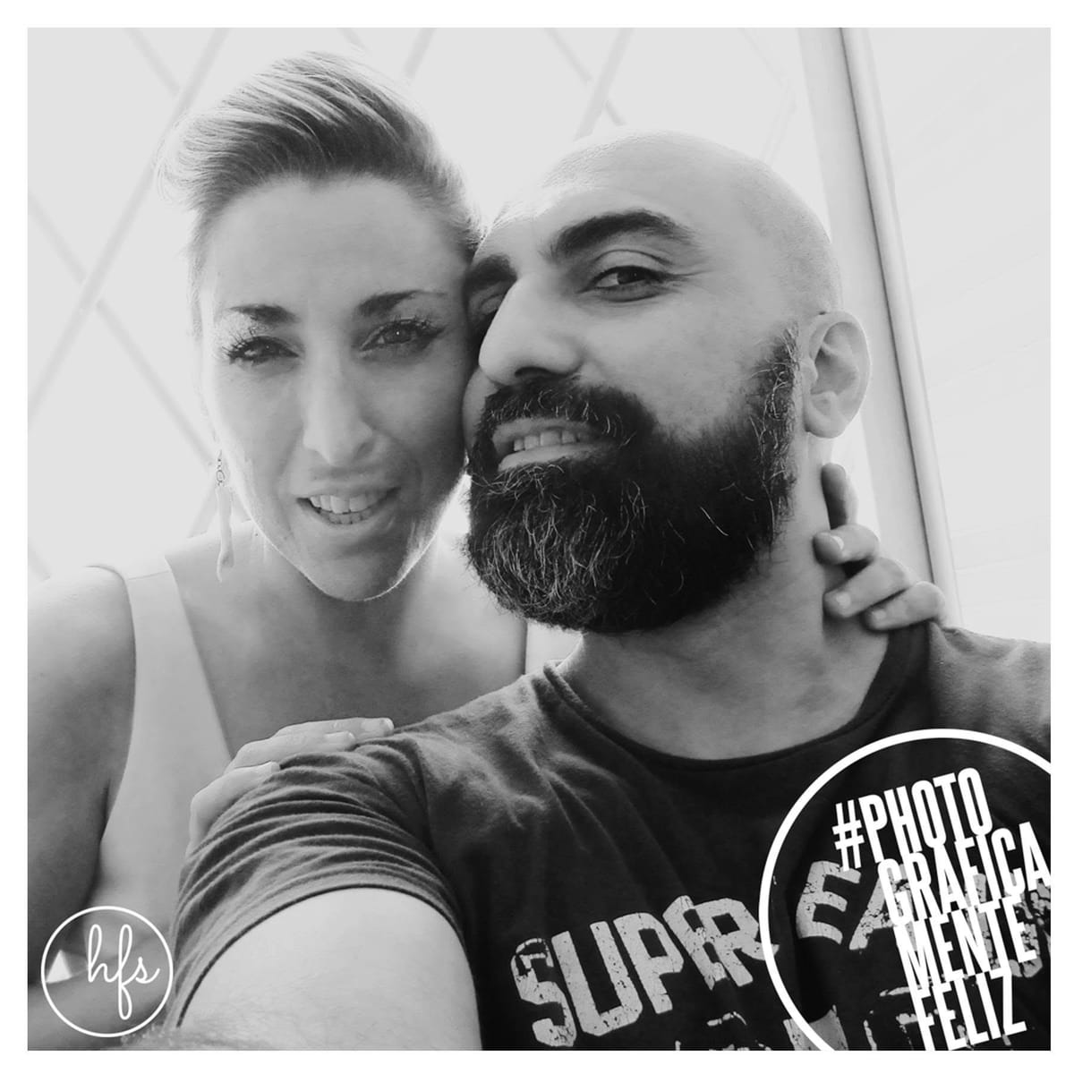 Selfie en blanco y negro de una pareja sonriente en un espacio con paredes tipo carpa o iglú, ella con corte pixie apoyando la cabeza en él, él calvo y con barba abrazándola con una mano al…