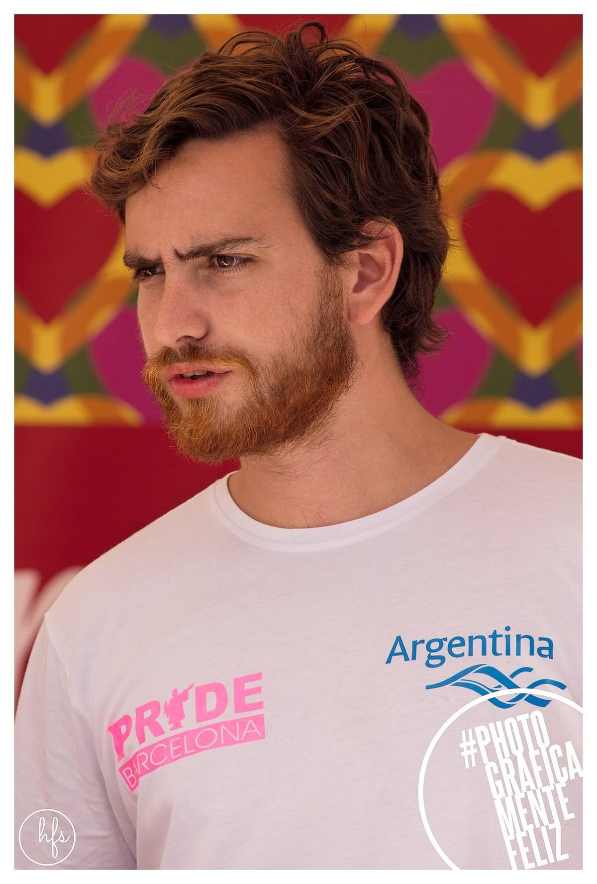 Retrato a color de un hombre de barba y pelo pelirrojos, con expresión de sorpresa o desconcierto, vistiendo una remera blanca con el logo "PRIDE BARCELONA" y "Argentina"; fondo decorativo…
