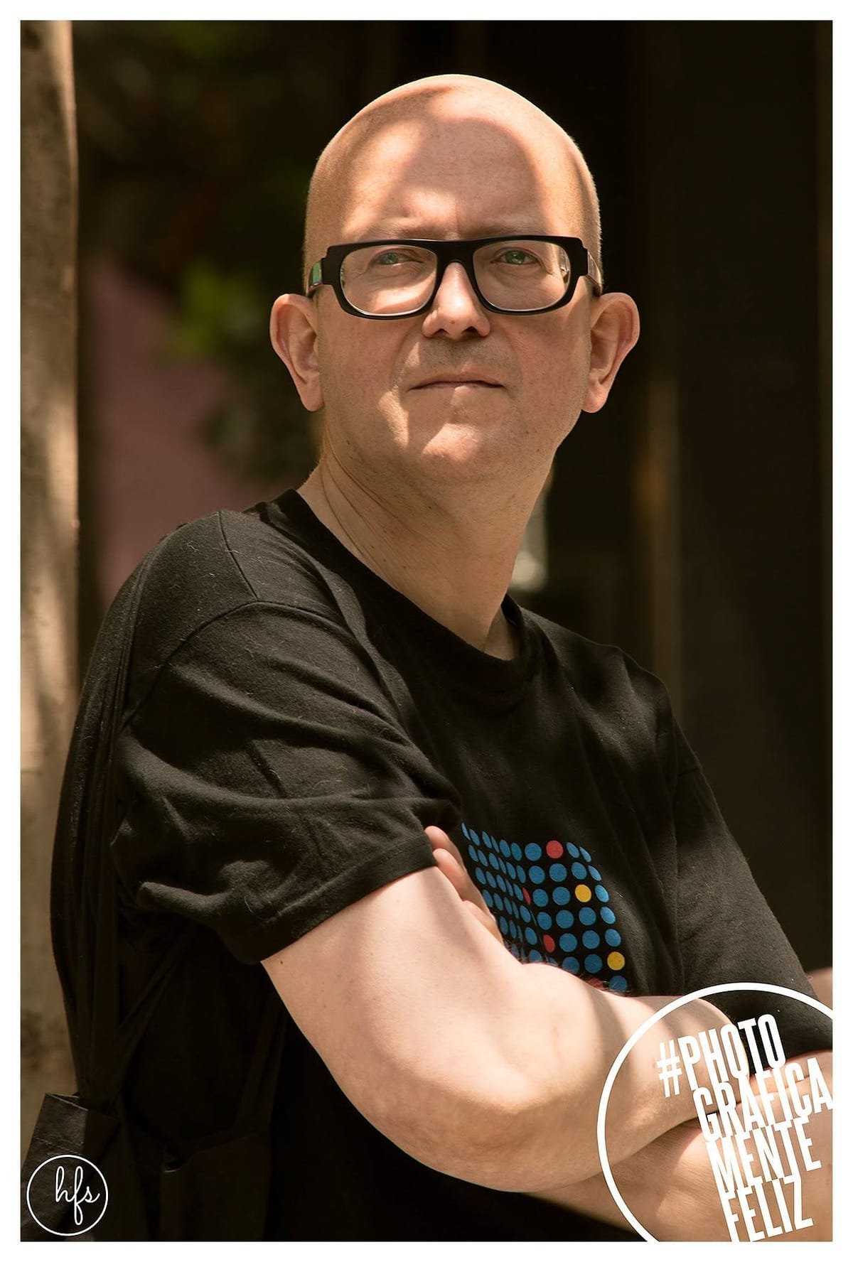 Retrato de un hombre calvo con anteojos de marco grueso negro, remera oscura con estampa de puntos de colores, brazos cruzados, posando frente a un portón de madera con luz cálida de…