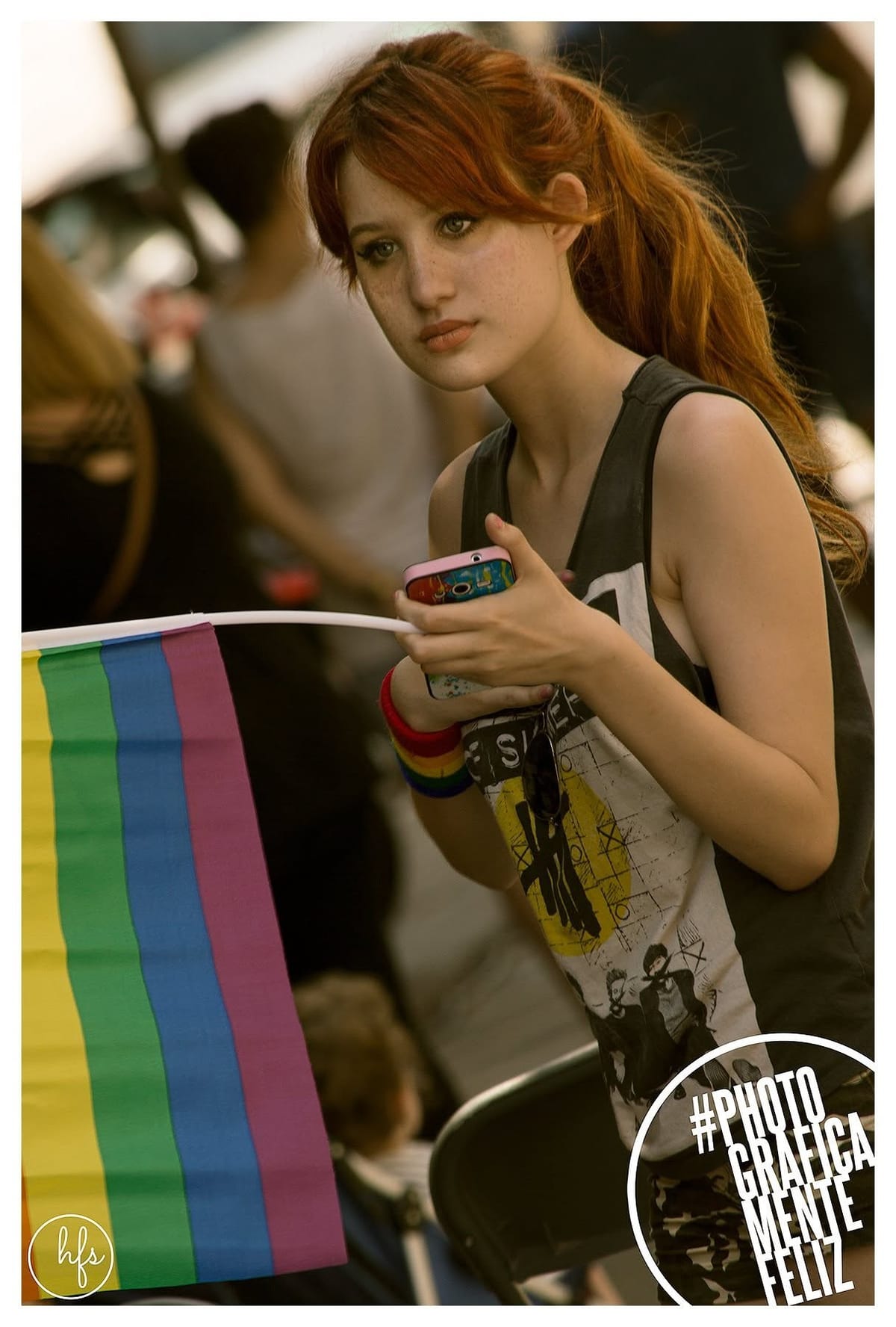 Retrato a color de una joven pelirroja con pecas, sosteniendo un teléfono y una bandera arcoíris con una pulsera del mismo motivo en la muñeca; viste una musculosa oscura estampada, con…