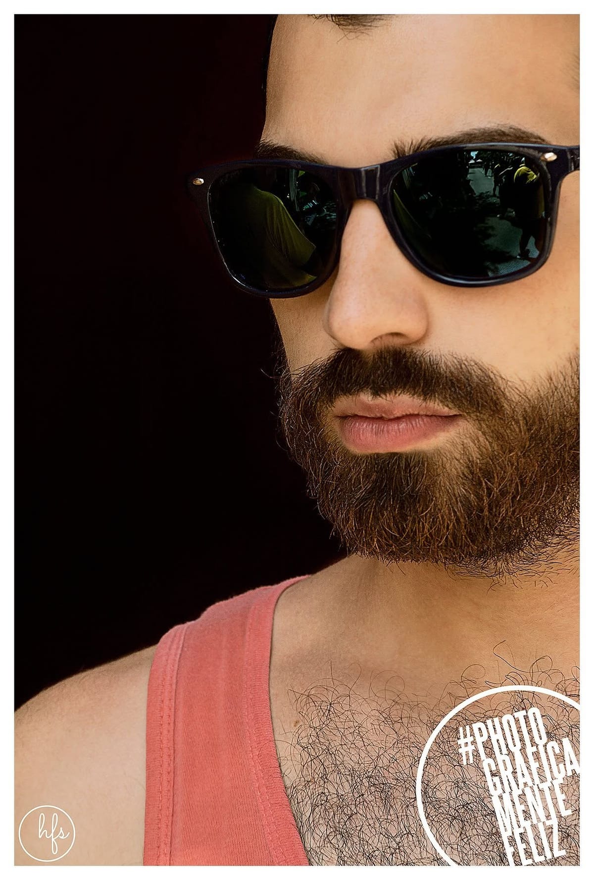 Primer plano muy cerrado de un hombre con anteojos de sol oscuros, barba prolija y musculosa rosa, iluminado por luz dura lateral que marca fuerte contraste entre sombra y piel.