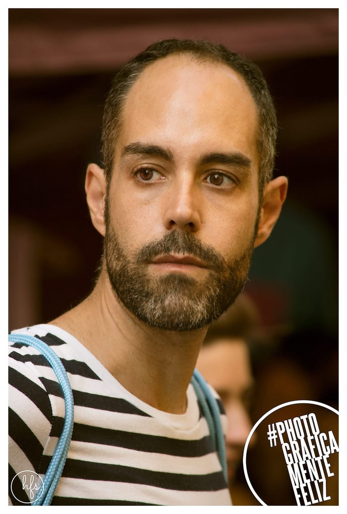 Retrato a color de un hombre de entradas pronunciadas y barba entrecana, con remera a rayas y un cordón celeste al cuello, mirando hacia un lado con expresión seria; fondo interior…