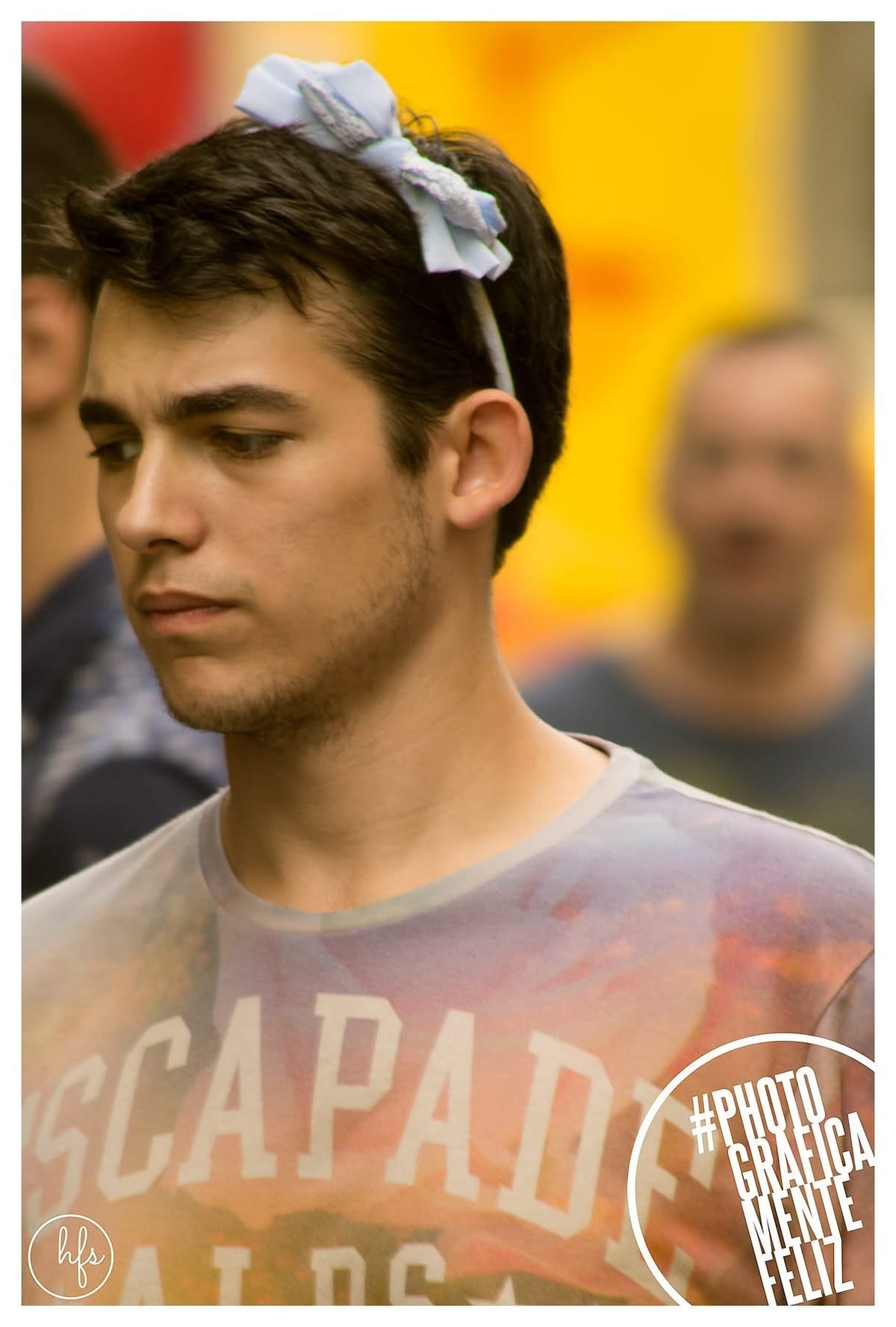 Retrato callejero de un joven de perfil con un llamativo moño de tela celeste sobre el pelo oscuro, remera con estampado desteñido de la palabra «Escapade».