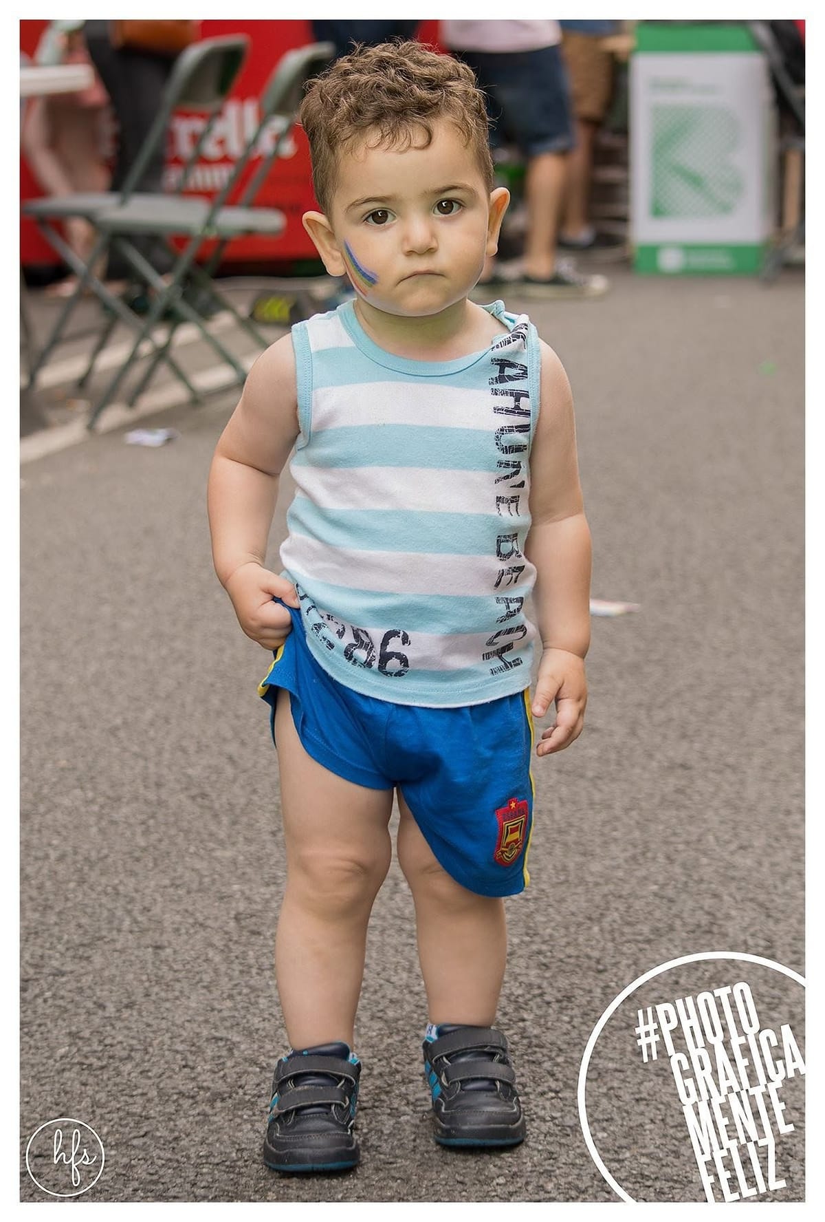 Retrato de cuerpo entero de un niño pequeño con musculosa a rayas celestes y short azul, mirando serio a cámara con una franja de colores tipo arcoíris pintada en la mejilla, de pie en una…