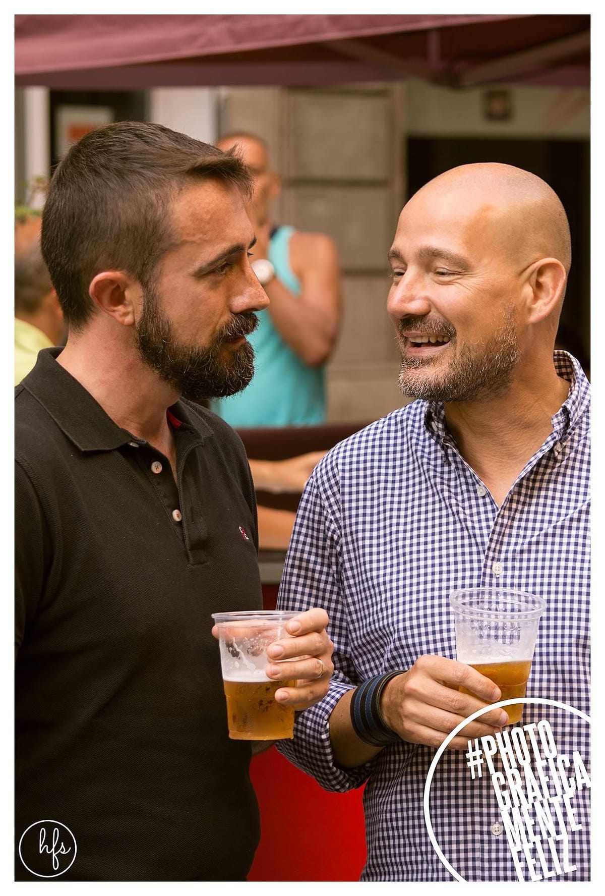 Retrato en color de dos hombres de mediana edad conversando y riendo, cada uno con un vaso de cerveza en la mano, bajo un toldo en una terraza; uno con barba oscura y remera negra, el otro…