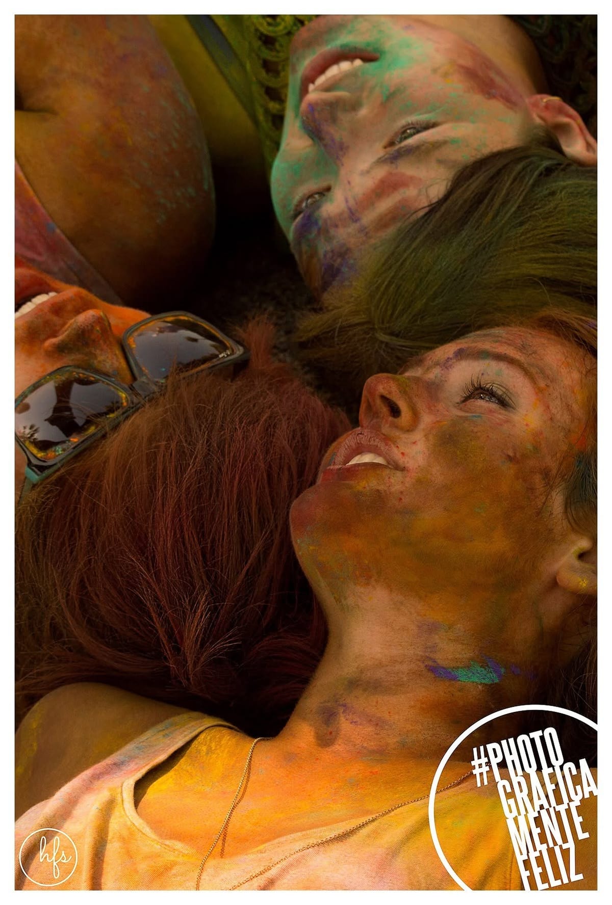 Tres mujeres recostadas boca arriba, rostros cubiertos de pigmentos de colores, riendo y mirándose entre ellas en un momento de complicidad.
