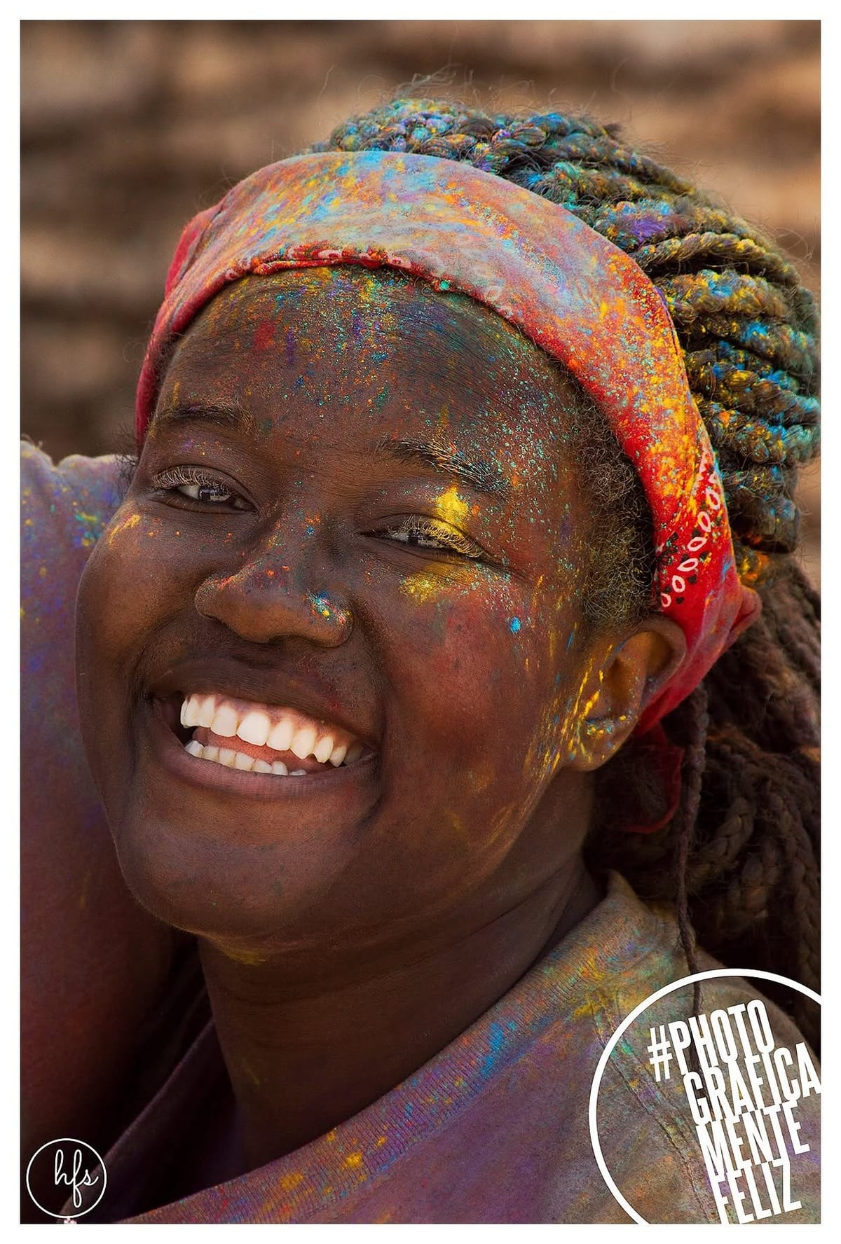 Retrato de una mujer con trenzas largas y un pañuelo rojo estampado como vincha, riendo ampliamente, con el rostro y las trenzas salpicados de pigmentos de colores propios de la fiesta Holi.