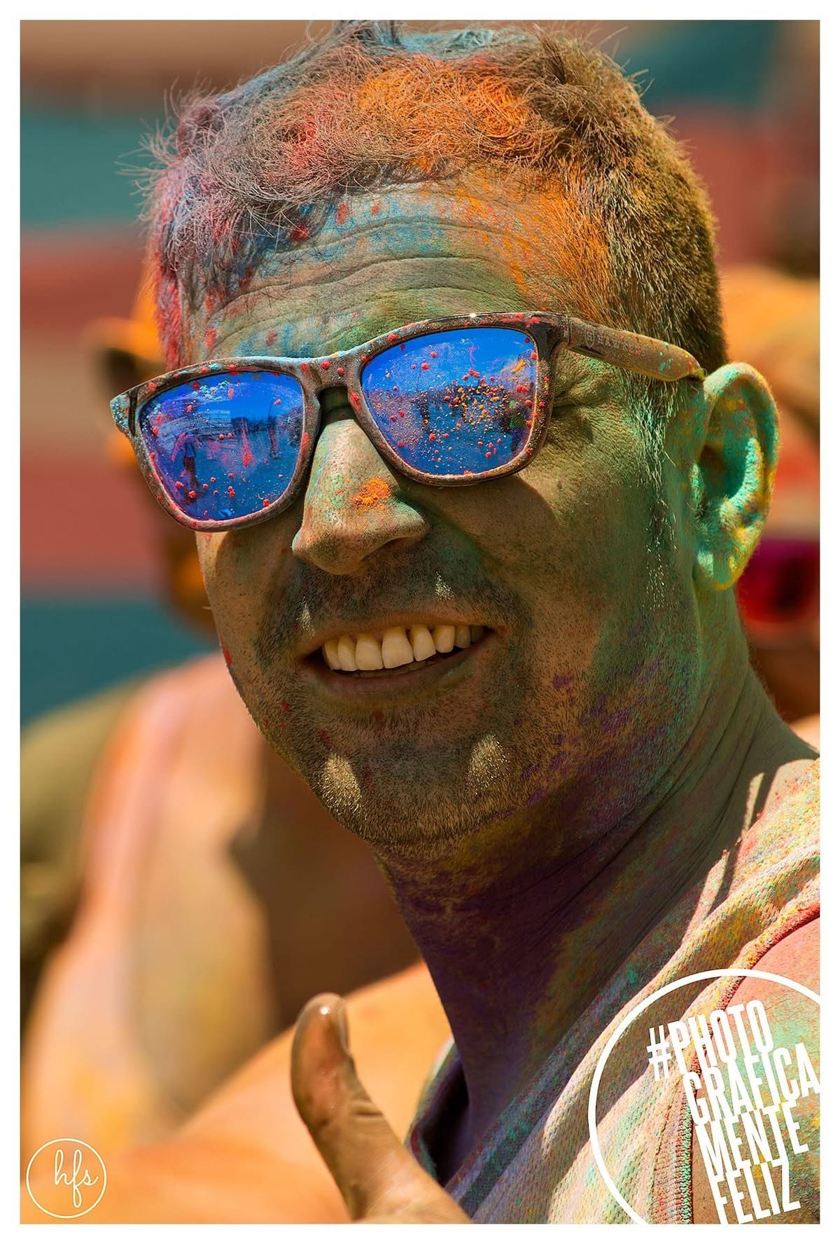 Primer plano de un hombre sonriente con anteojos de sol reflectantes azules donde se refleja el entorno festivo, rostro y pelo cubiertos de pigmentos verde, naranja y celeste propios de la…