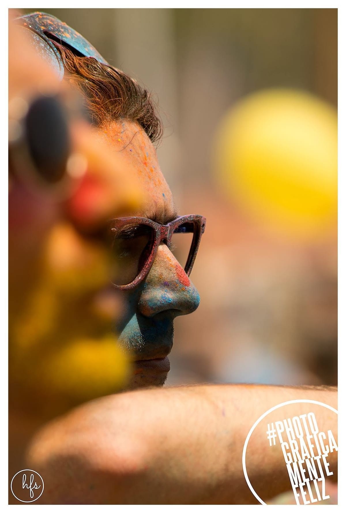 Retrato de perfil a color de un hombre con gafas de sol y el rostro cubierto de polvo Holi azul y naranja, con mechones de pelo también teñidos; al fondo, una gran esfera amarilla…