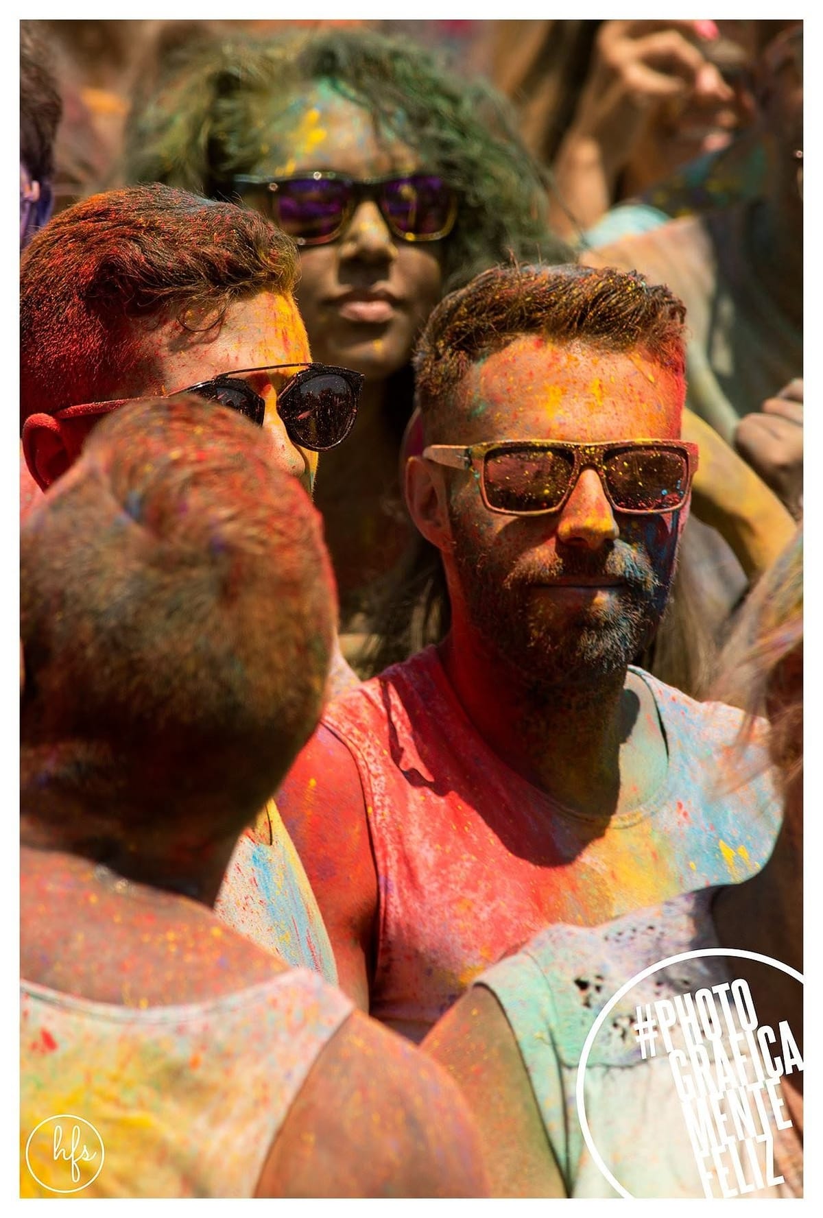 Grupo de tres personas cubiertas de polvo de colores Holi: en primer plano dos hombres con anteojos de sol oscuros se miran de perfil, mientras una tercera persona con anteojos violeta…