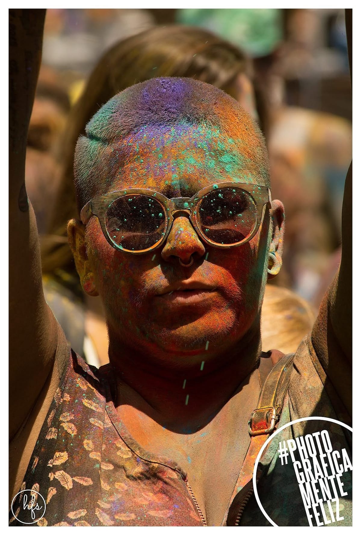 Retrato frontal de una persona con la cabeza rapada cubierta de polvo turquesa y naranja, gafas redondas puestas sobre el rostro y un piercing en la nariz, con expresión seria y boca…