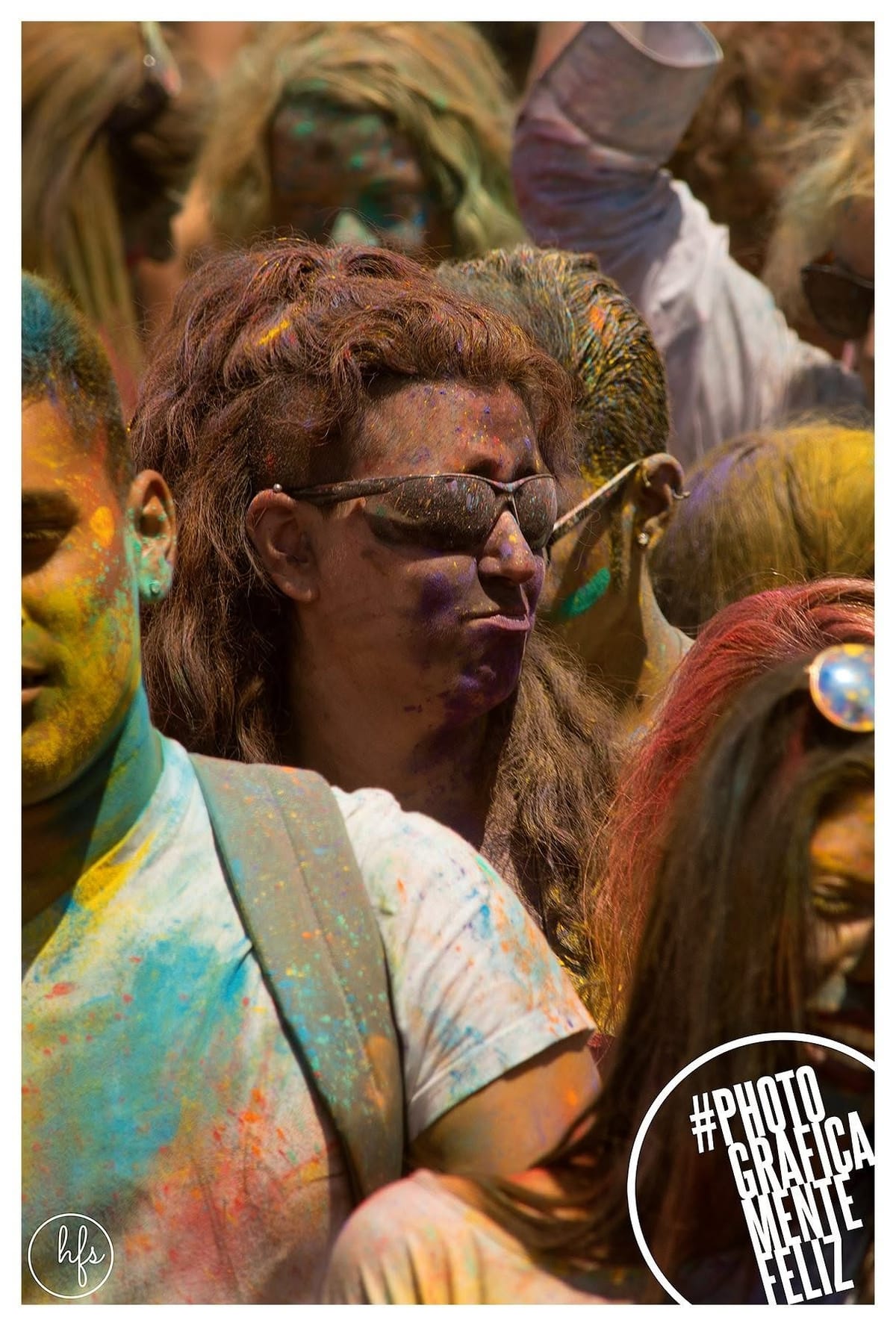 Retrato de una persona de melena castaña ondulada y anteojos de sol oscuros, rostro cubierto de pigmentos violeta y azul, rodeada muy de cerca por otras caras también pintadas de la fiesta…