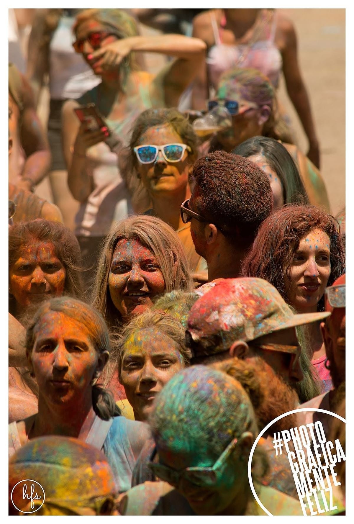 Fotografía a color de una multitud apretada de jóvenes con el rostro y el pelo cubiertos de polvo Holi en múltiples colores, mirando en distintas direcciones; algunos llevan gafas de sol y…