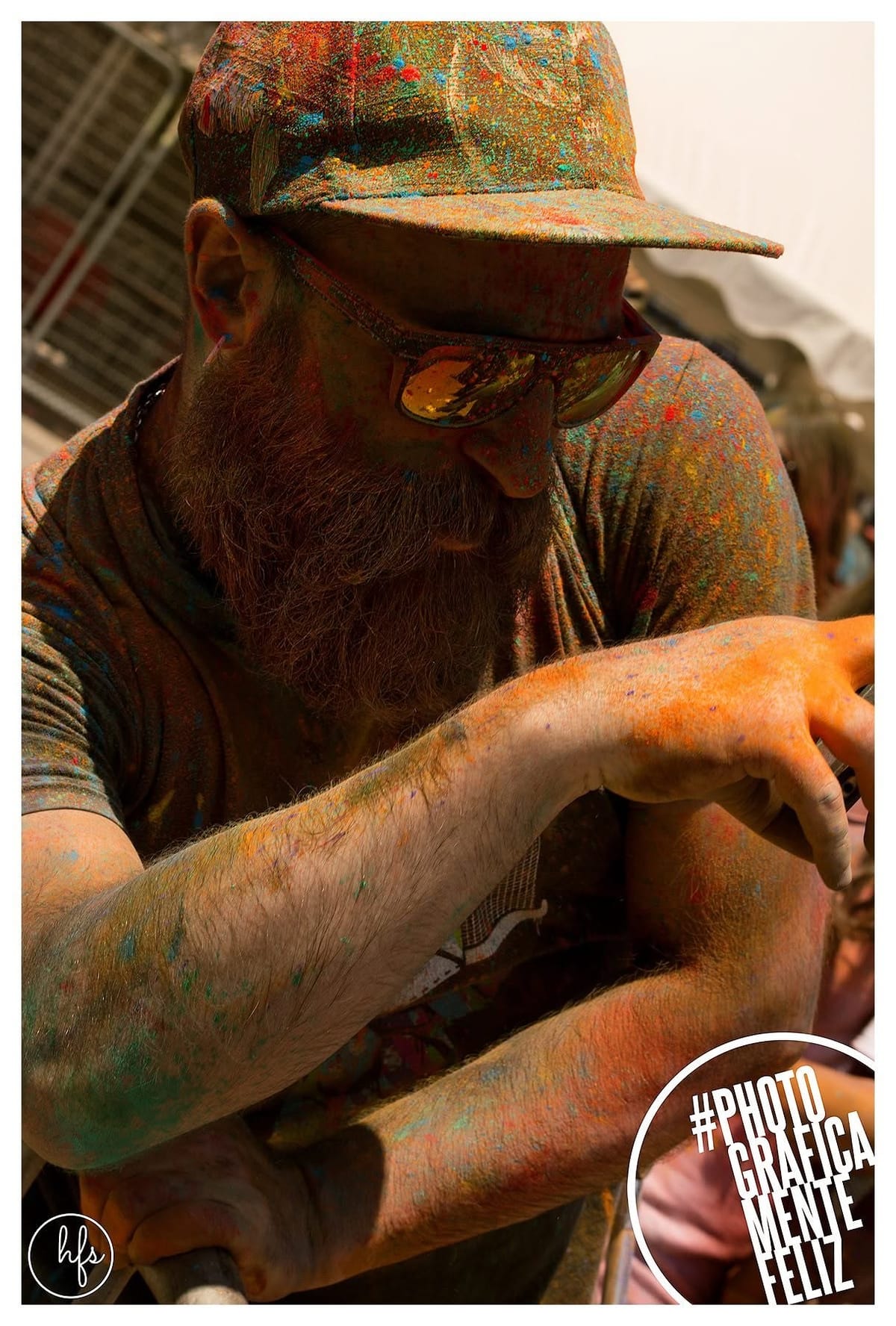 Hombre con barba tupida y gorra, anteojos de sol, cubierto de pigmentos de colores propios del festival Holi en rostro, brazos y ropa.