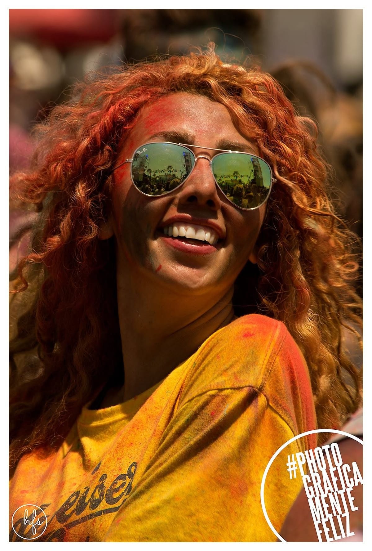 Retrato de una joven de pelo rizado pelirrojo con la piel cubierta de polvo naranja y rosa, gafas de sol espejadas que reflejan la calle, riendo con la cabeza inclinada hacia atrás bajo luz…