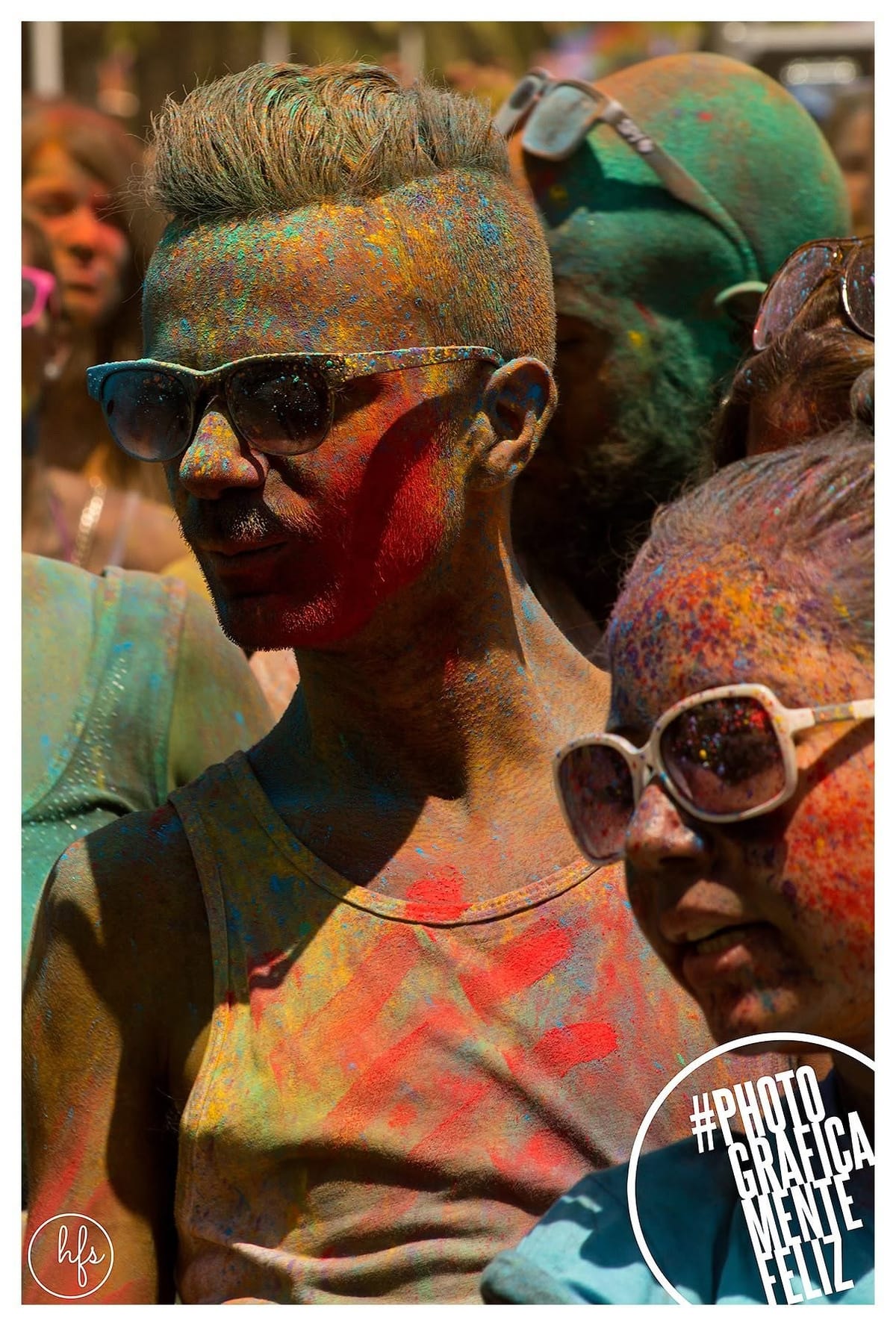 Retrato de un hombre con anteojos de sol y mitad del rostro cubierta de pintura roja, cabello teñido de rubio, entre otras personas también pintadas de la fiesta Holi.
