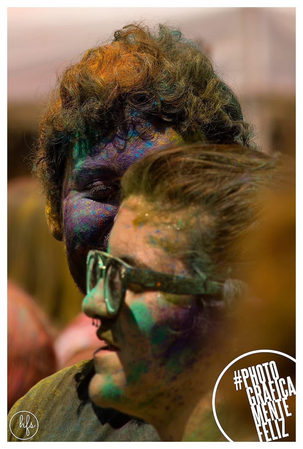 Composición a color de dos personas con el rostro cubierto de pigmentos Holi en tonos azules, violetas y dorados; una de ellas usa anteojos y aparece en primer plano mirando hacia abajo…