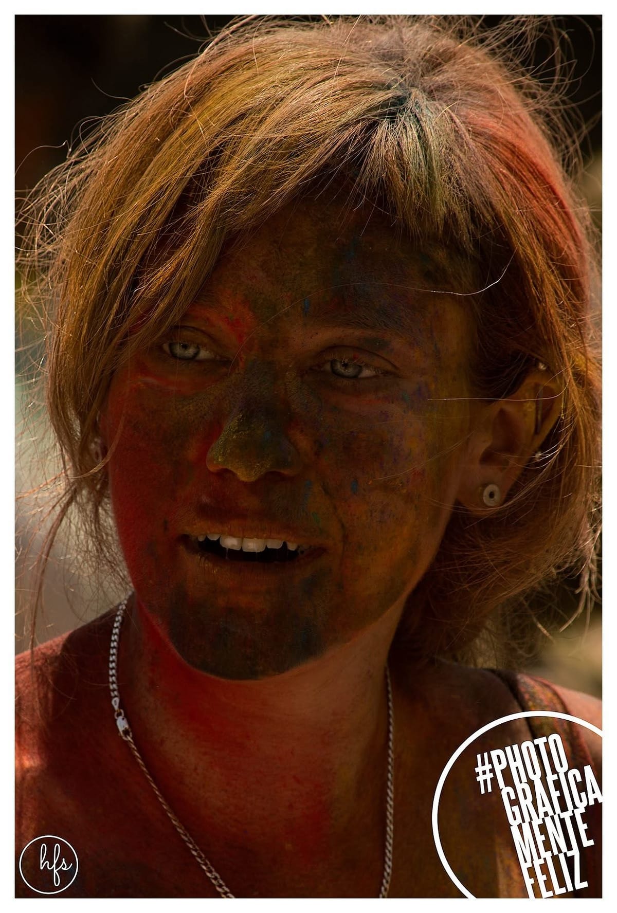 Retrato de una persona con el pelo rubio corto cubierto de tinte multicolor y la cara pintada de rojo y naranja de forma irregular, mirando fijo a cámara con expresión seria, luz cálida de…
