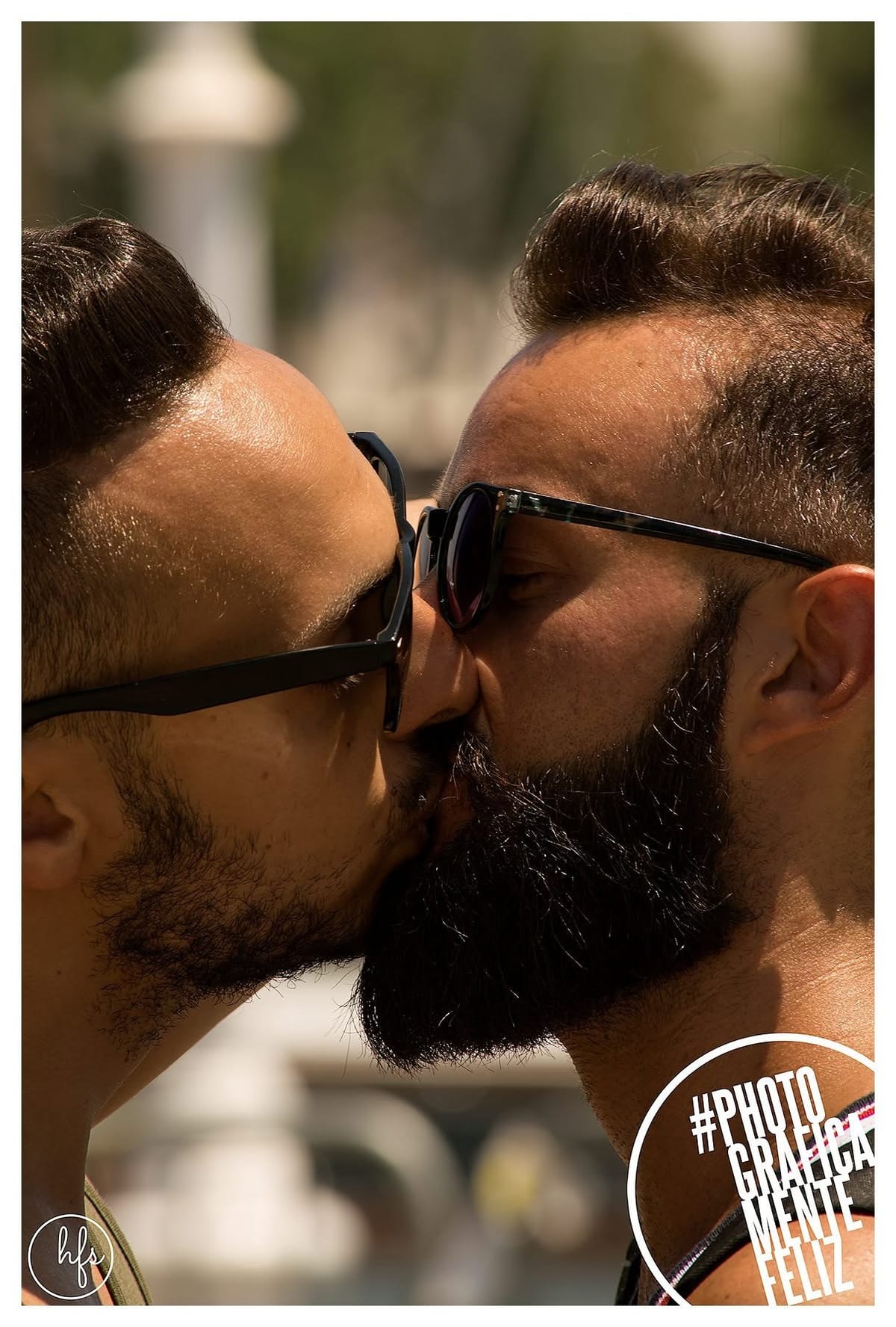 Primer plano de dos hombres con gafas de sol besándose, frentes y narices casi tocándose, con barba abundante uno de ellos; fondo urbano desenfocado bajo luz de sol intensa.