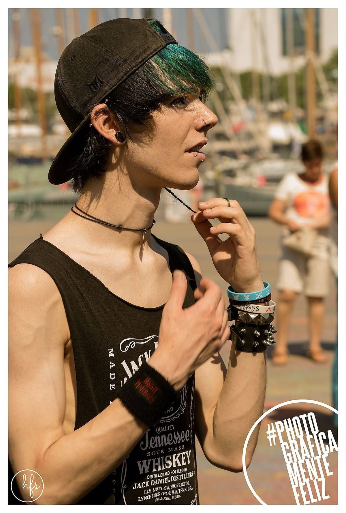 Retrato de perfil de un joven con gorra hacia atrás y mechón teñido de verde azulado, múltiples piercings en oreja y septum, con gargantilla de cuero y pulseras, fondo de un puerto…