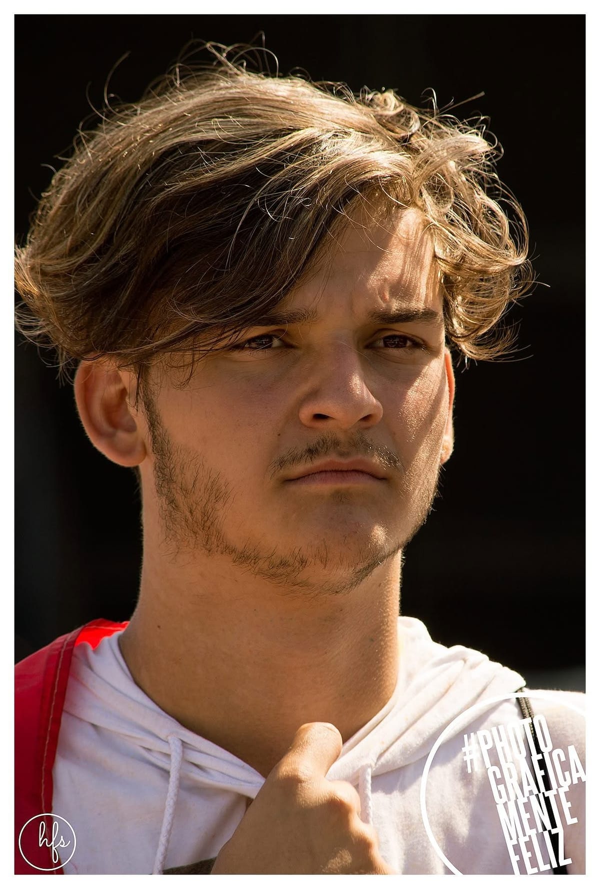Retrato a color de un joven de pelo castaño despeinado y mirada seria, iluminado por luz solar rasante que marca fuertes contrastes en el rostro; viste una prenda blanca con un tirante rojo…