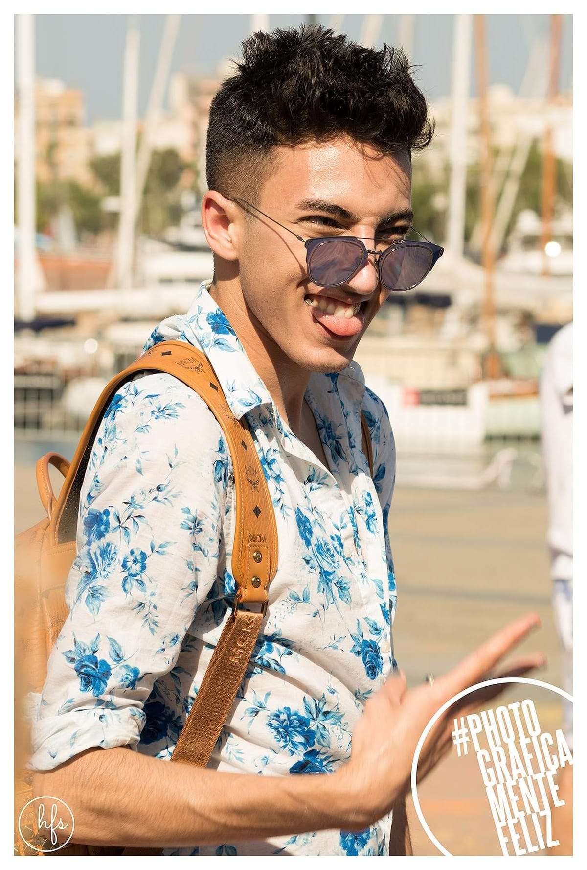 Retrato a color de un joven de pelo oscuro peinado hacia arriba, con gafas de sol y una mochila color camel, sacando la lengua y riendo mientras hace un gesto con la mano; al fondo…