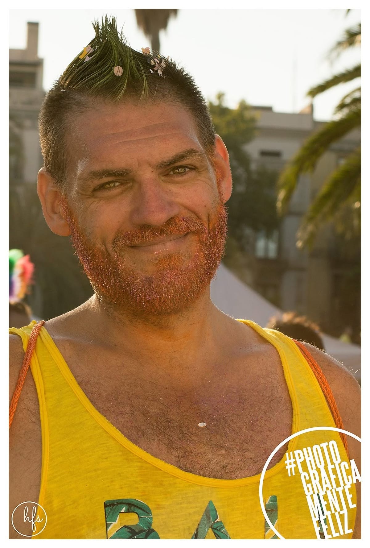 Retrato de un hombre con cresta pintada de verde con confeti pegado, barba pelirroja con brillantina rosa y camiseta amarilla, sonriendo de frente con un ambiente urbano desenfocado de fondo.