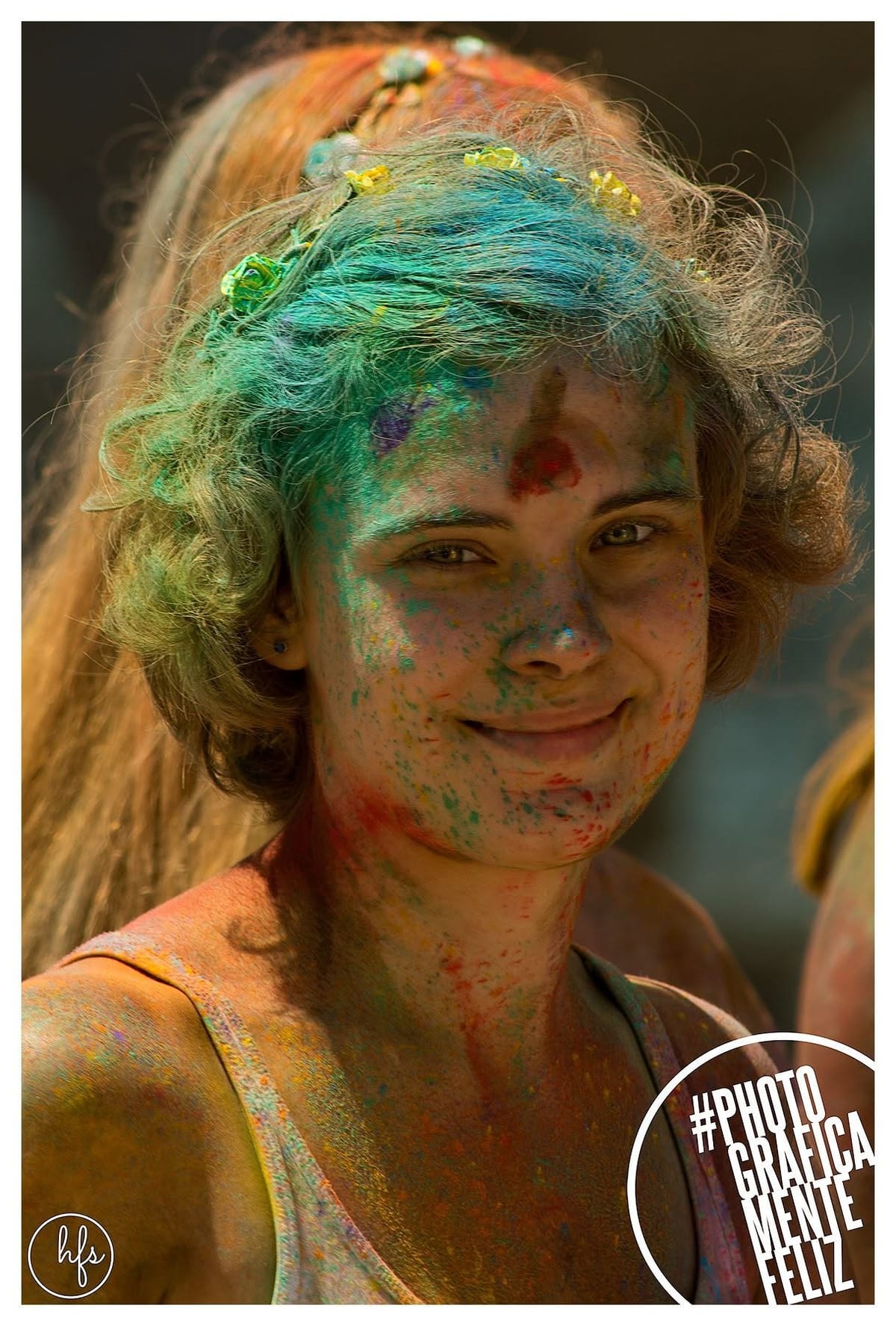 Retrato de una joven con el pelo teñido de verde y azul por el polvo de colores, corona de flores, y la cara salpicada de pigmentos rojo, verde y amarillo, sonriendo de frente bajo luz…
