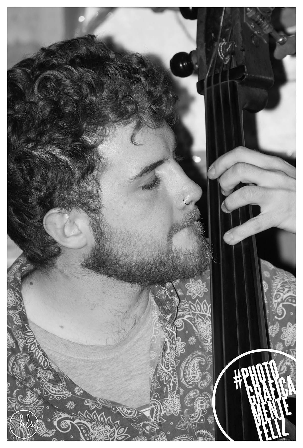 Retrato en blanco y negro de un músico de perfil, pelo rizado y aro en la nariz, con los ojos cerrados apoyando la mejilla casi sobre el mástil del contrabajo mientras su mano pulsa las…