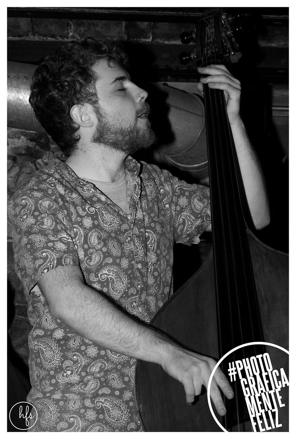 Retrato en blanco y negro de un músico de perfil, ojos cerrados y boca entreabierta cantando, con camisa estampada de cachemira, tocando un contrabajo bajo luz ambiente de bar; se distingue…