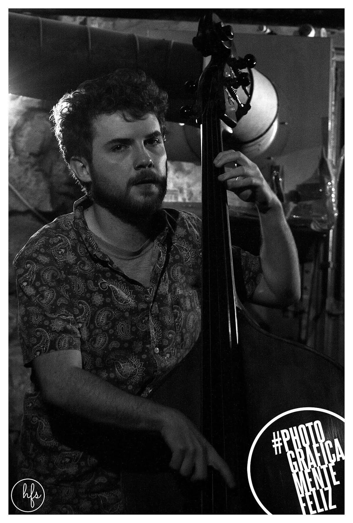 Retrato en blanco y negro de un músico de rulos oscuros y barba, con camisa estampada de cachemira, tocando un contrabajo y mirando directamente a cámara; fondo de pared de piedra con foco…