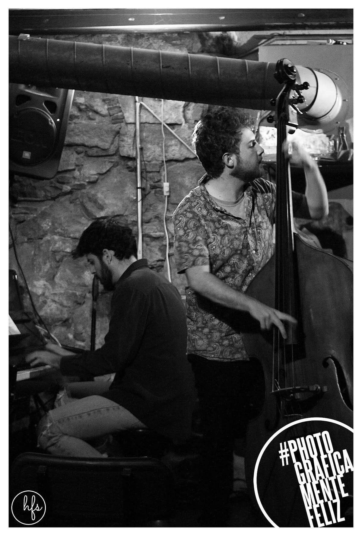 Foto en blanco y negro de una banda de jazz tocando en un local de piedra vista, con un contrabajista de pie iluminado por un foco cenital, un pianista sentado a la izquierda y un tercer…