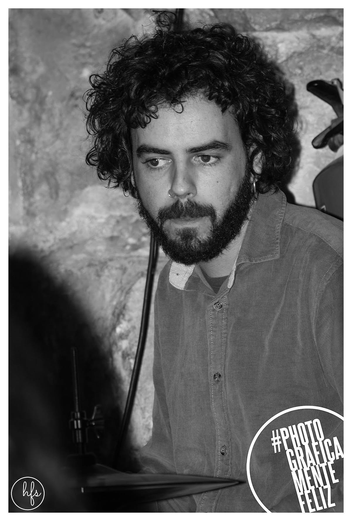 Retrato en blanco y negro de un baterista de cabello rizado y barba, con piercing en el septum, mirando hacia abajo mientras toca; se ve parte del platillo y el pie del micrófono, fondo de…