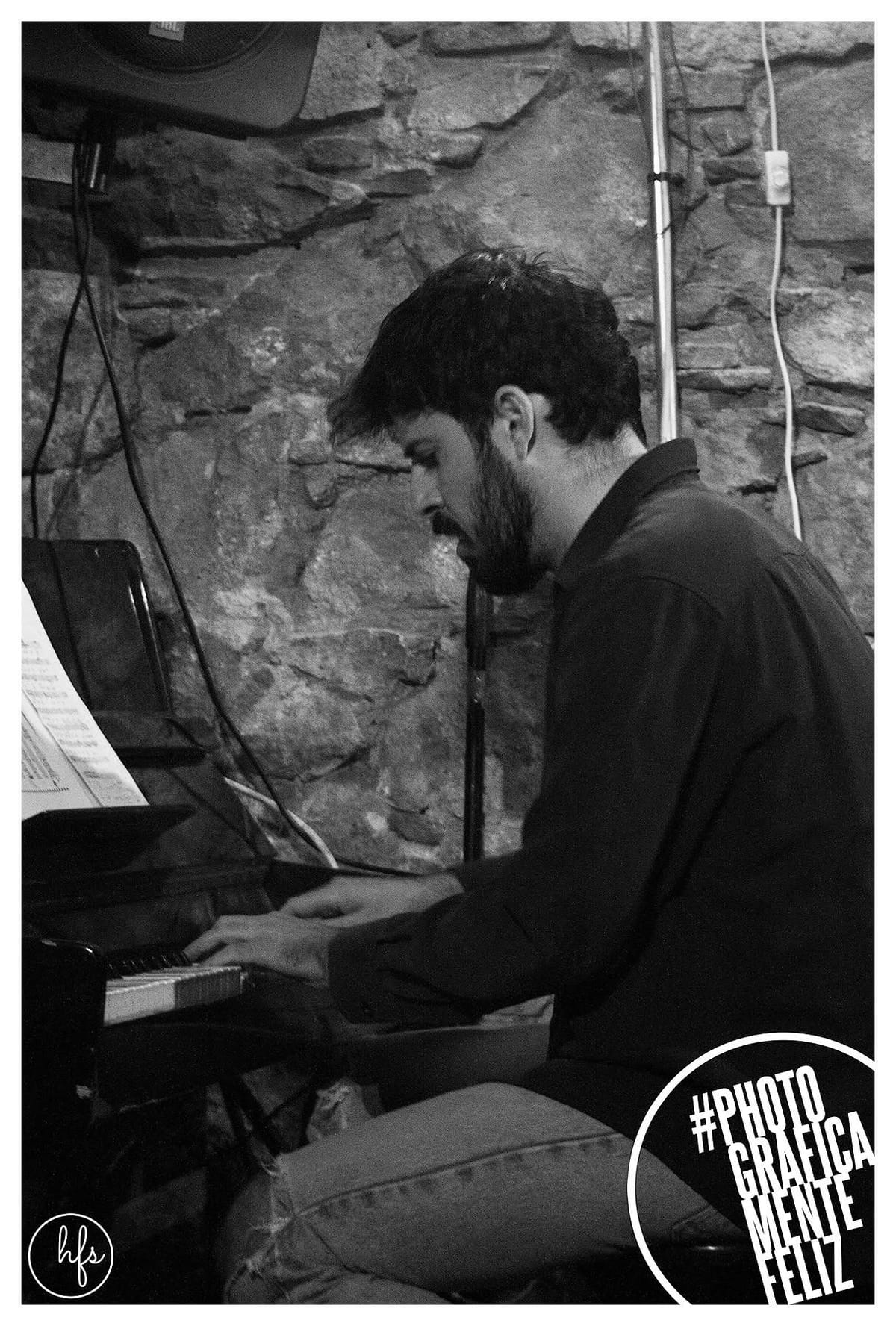 Fotografía en blanco y negro de un pianista de perfil, cabello oscuro despeinado y barba, tocando un piano de cola ante una partitura, con un muro de piedra vista detrás y cables e…