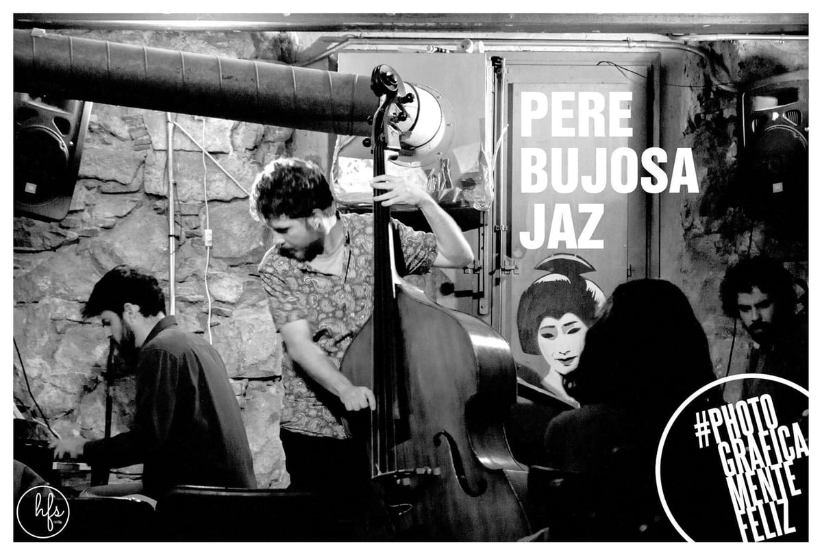 Fotografía en blanco y negro de un trío de jazz tocando en un local de paredes de piedra vista; en primer plano un contrabajista con camisa estampada apoya el instrumento junto a un cartel…