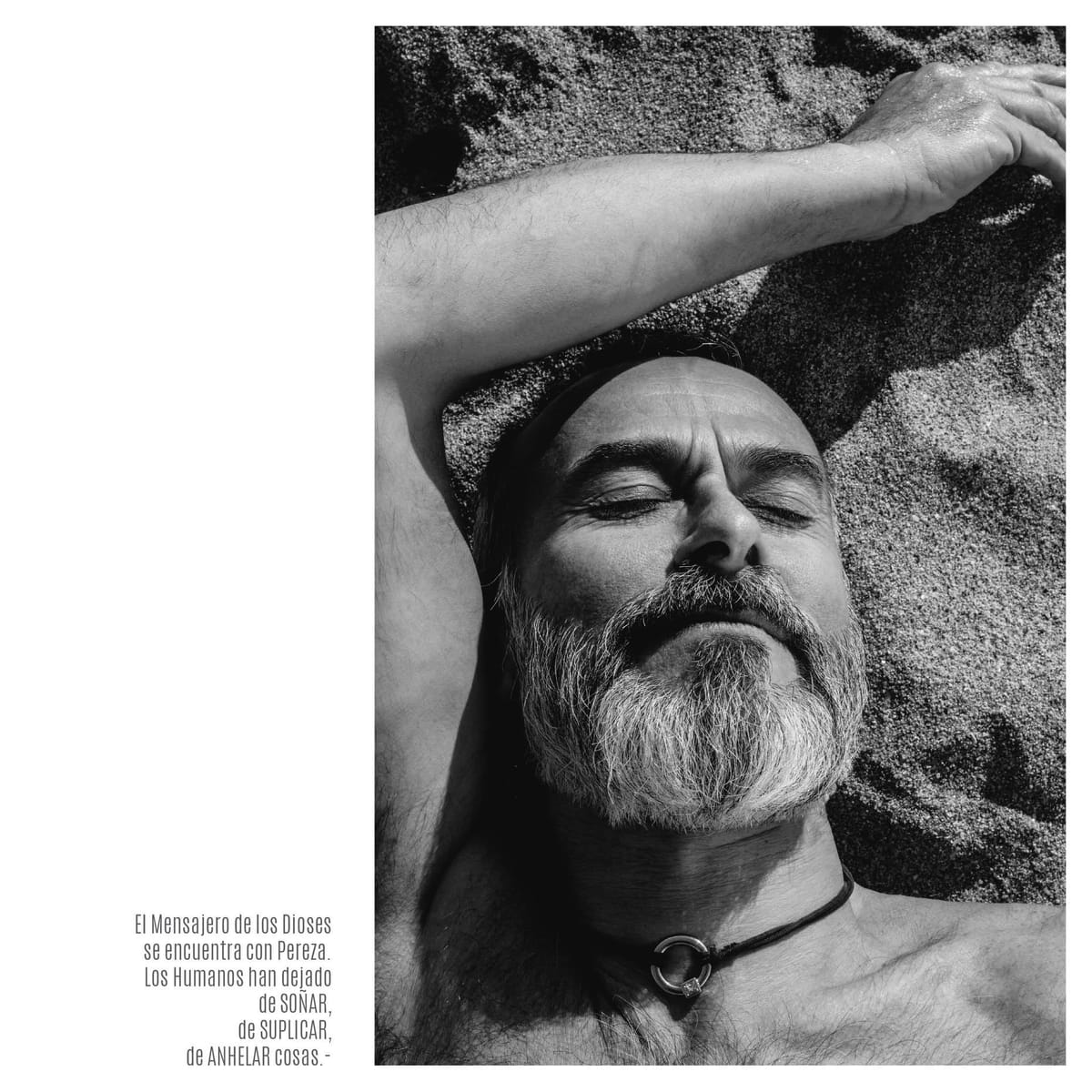Retrato en blanco y negro de un hombre calvo con barba y bigote gris, tendido boca arriba sobre arena gruesa, ojos cerrados y un brazo cruzado sobre la frente; lleva un collar de cuero con…