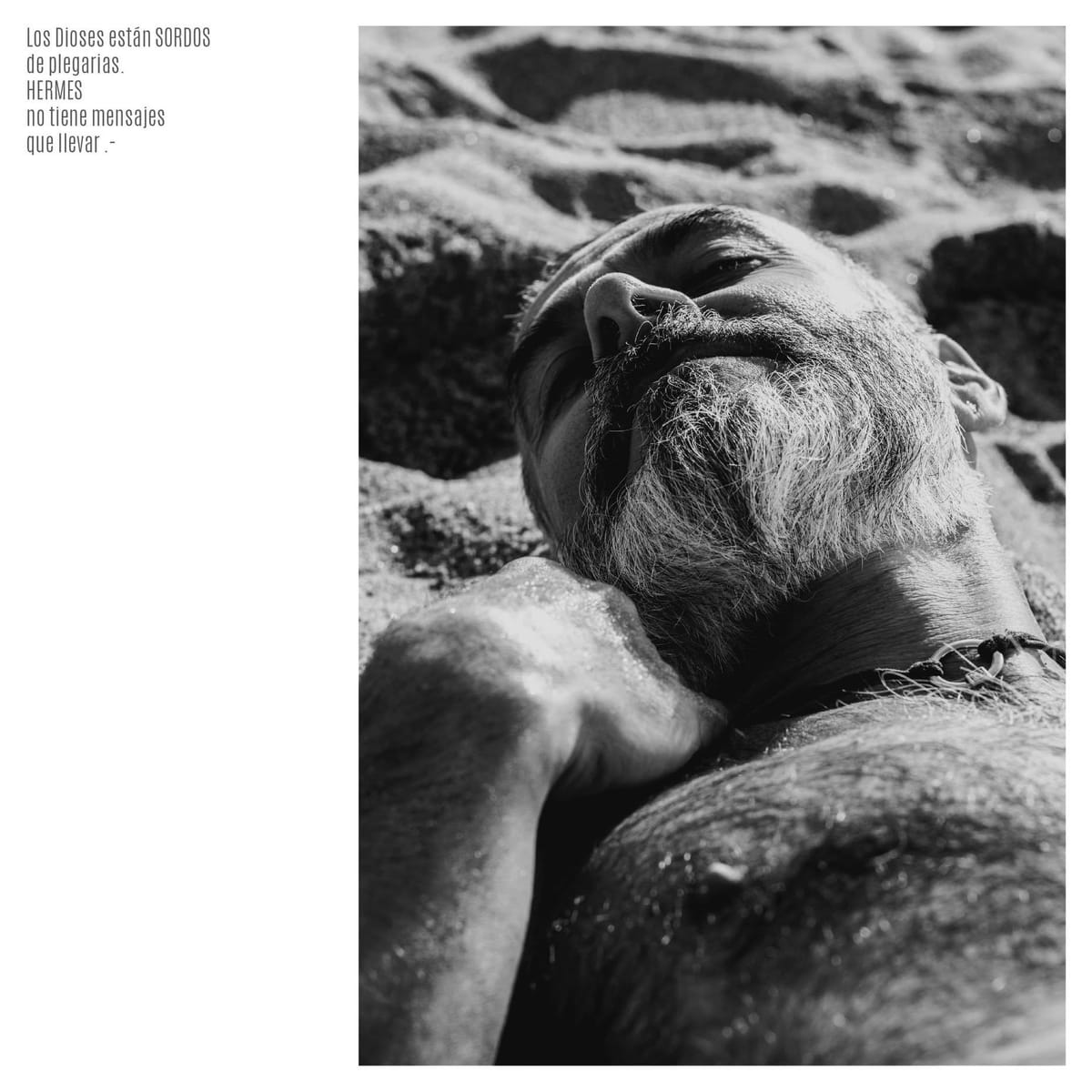 Fotografía en blanco y negro de un hombre mayor de barba blanca recostado sobre arena o piedra, con la cabeza echada hacia atrás y los ojos entrecerrados; un brazo cruza en primer plano.