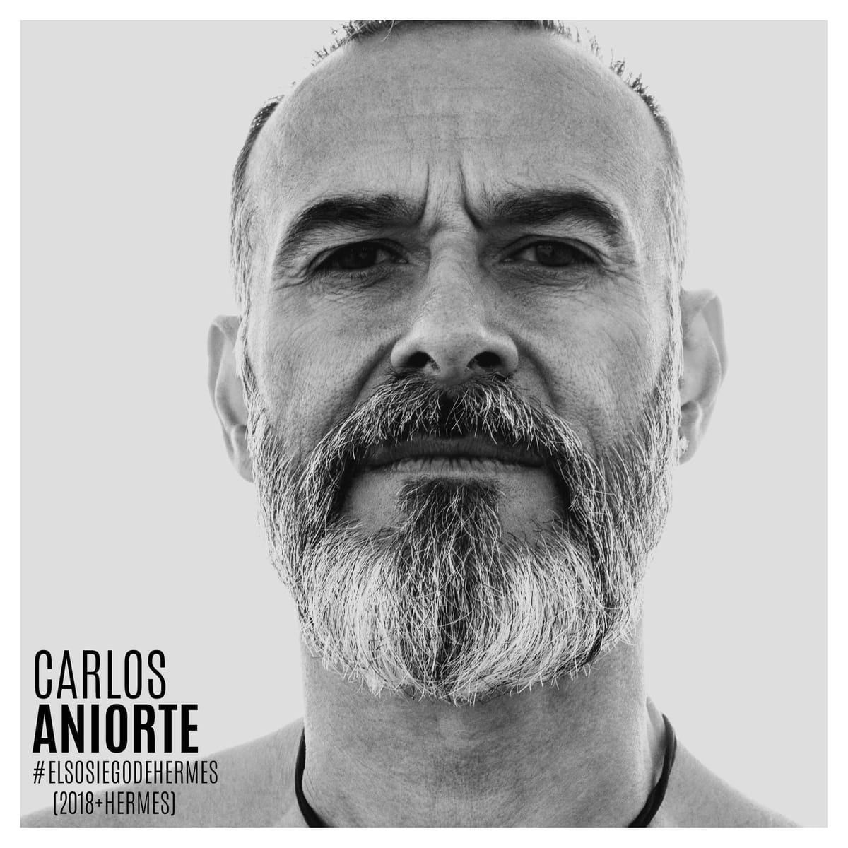 Retrato frontal en blanco y negro de un hombre calvo con barba entrecana larga, mirada fija a cámara sobre fondo gris liso.