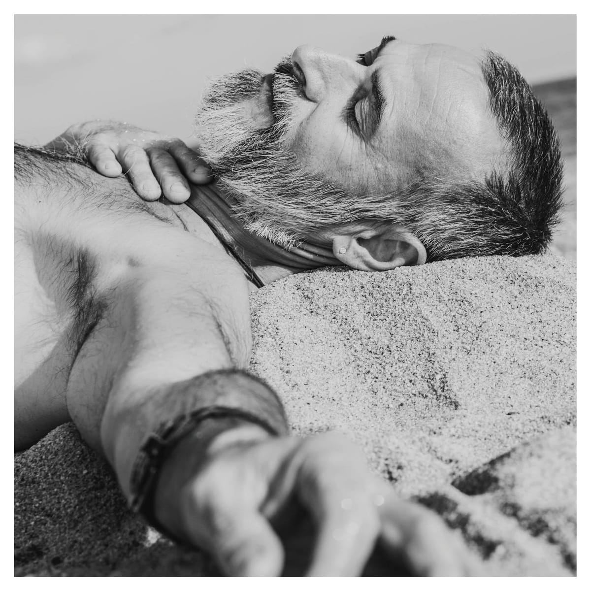 Retrato en blanco y negro de un hombre mayor de barba entrecana, torso desnudo y vello corporal denso, recostado en la playa con la cabeza apoyada sobre un montículo de arena y los ojos…