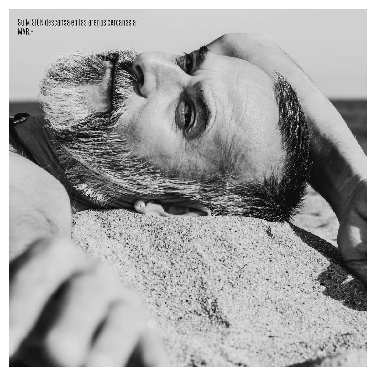 Segundo retrato en blanco y negro del mismo hombre, ahora de perfil, tendido con la cabeza hacia abajo sobre arena, la línea del mar visible al fondo.