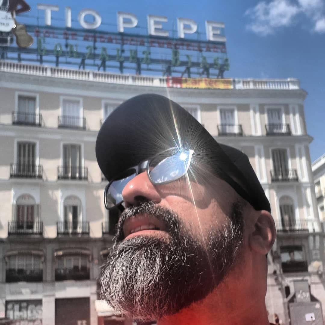 Autorretrato en contrapicado de un hombre con gorra, barba entrecana y anteojos de sol, tomado desde abajo con el cartel luminoso de «Tío Pepe» y la fachada de la Puerta del Sol de fondo.