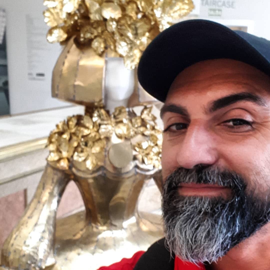 Selfie de un hombre con gorra oscura y barba entrecana, en primer plano ante una escultura dorada de una menina con volados y lazo, en un interior con paredes claras.