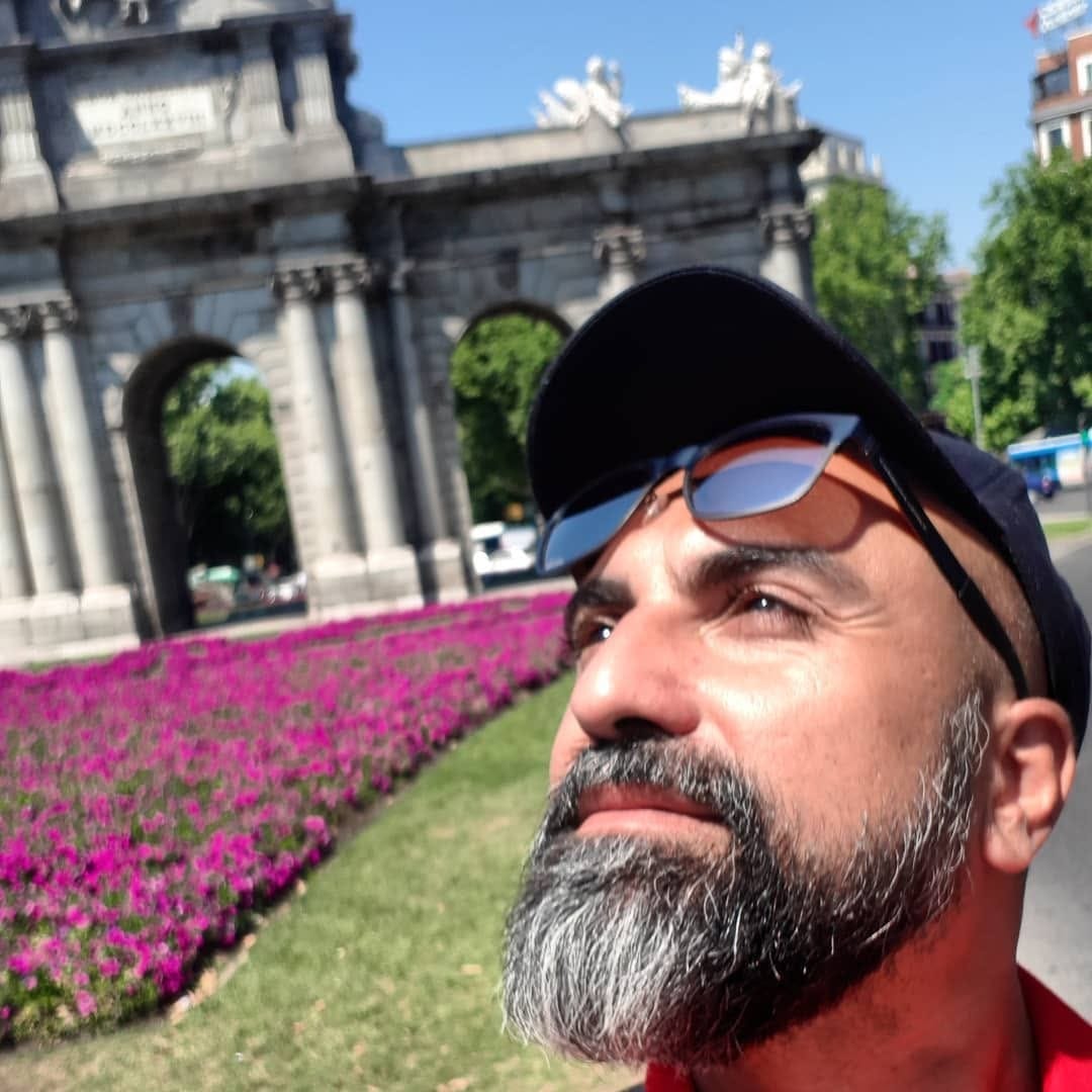 Selfie en primer plano de un hombre con barba entrecana, gafas de sol sobre una gorra oscura y remera roja, mirando hacia arriba bajo el sol.