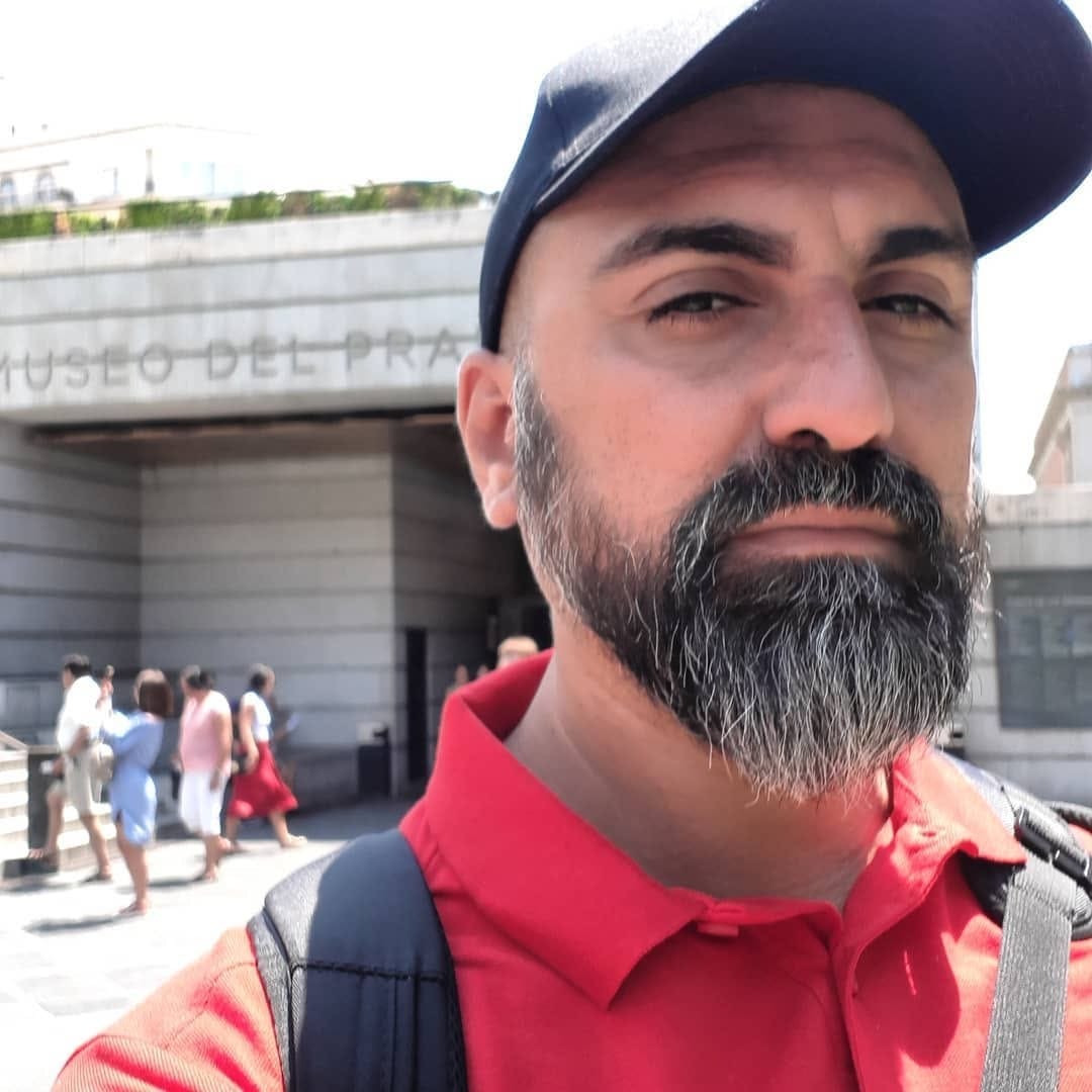 Selfie en primer plano de un hombre de barba entrecana con gorra negra y remera roja, mochila al hombro, con la fachada y el cartel "MUSEO DEL PRADO" detrás y transeúntes borrosos al fondo…