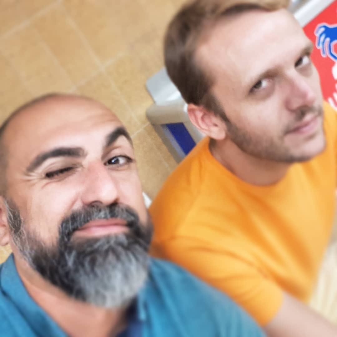 Selfie de gran angular con dos hombres: uno guiña un ojo en primer plano con barba entrecana, el otro con remera amarilla queda algo desenfocado detrás.